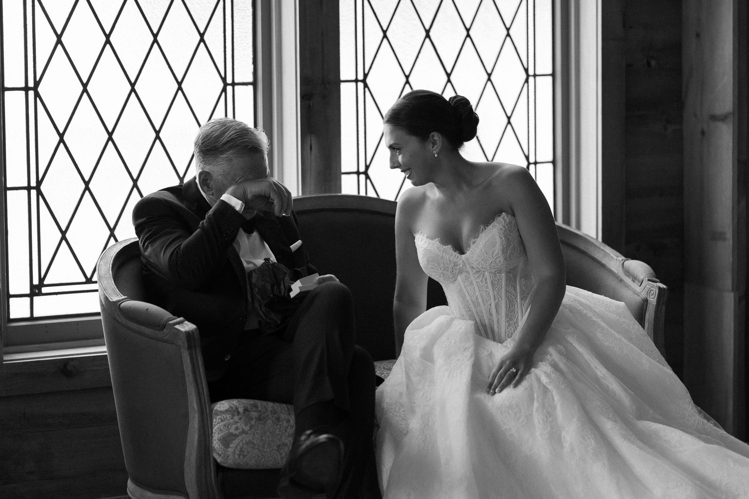 Black and white photo of emotional father wiping tears beside bride in wedding gown near leaded glass windows.
