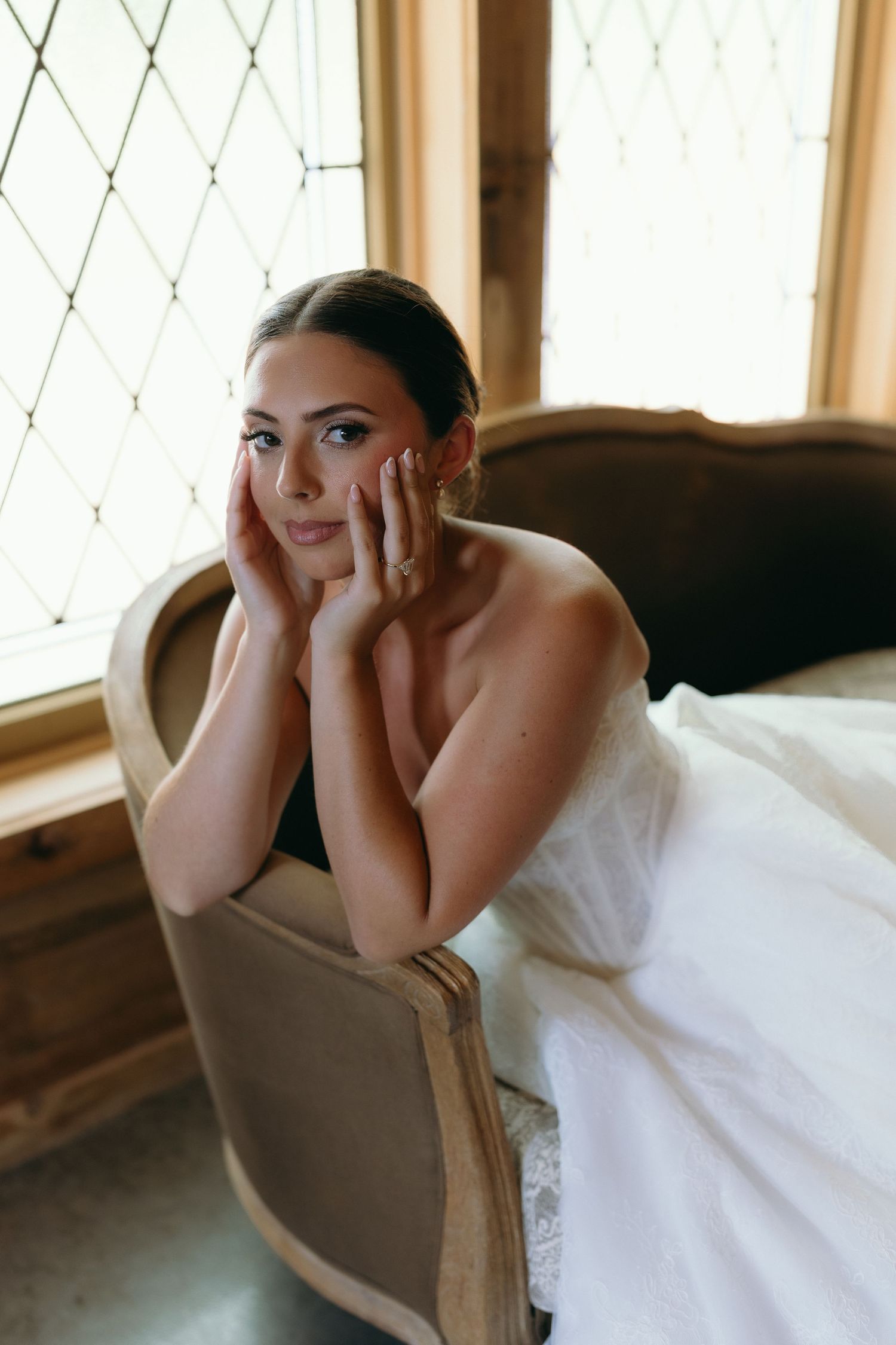 Elegant bride in strapless white gown sitting in vintage chair by diamond-paned window with hands on face.