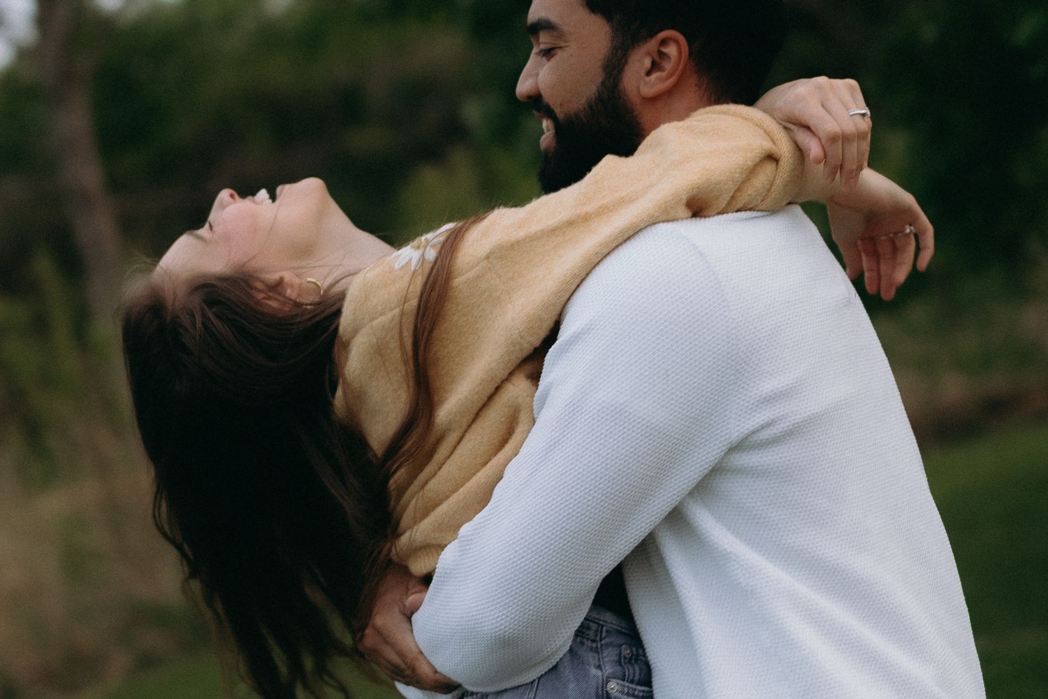A couple embracing outdoors, woman leaning back laughing joyfully while bearded man smiles, surrounded by green trees.