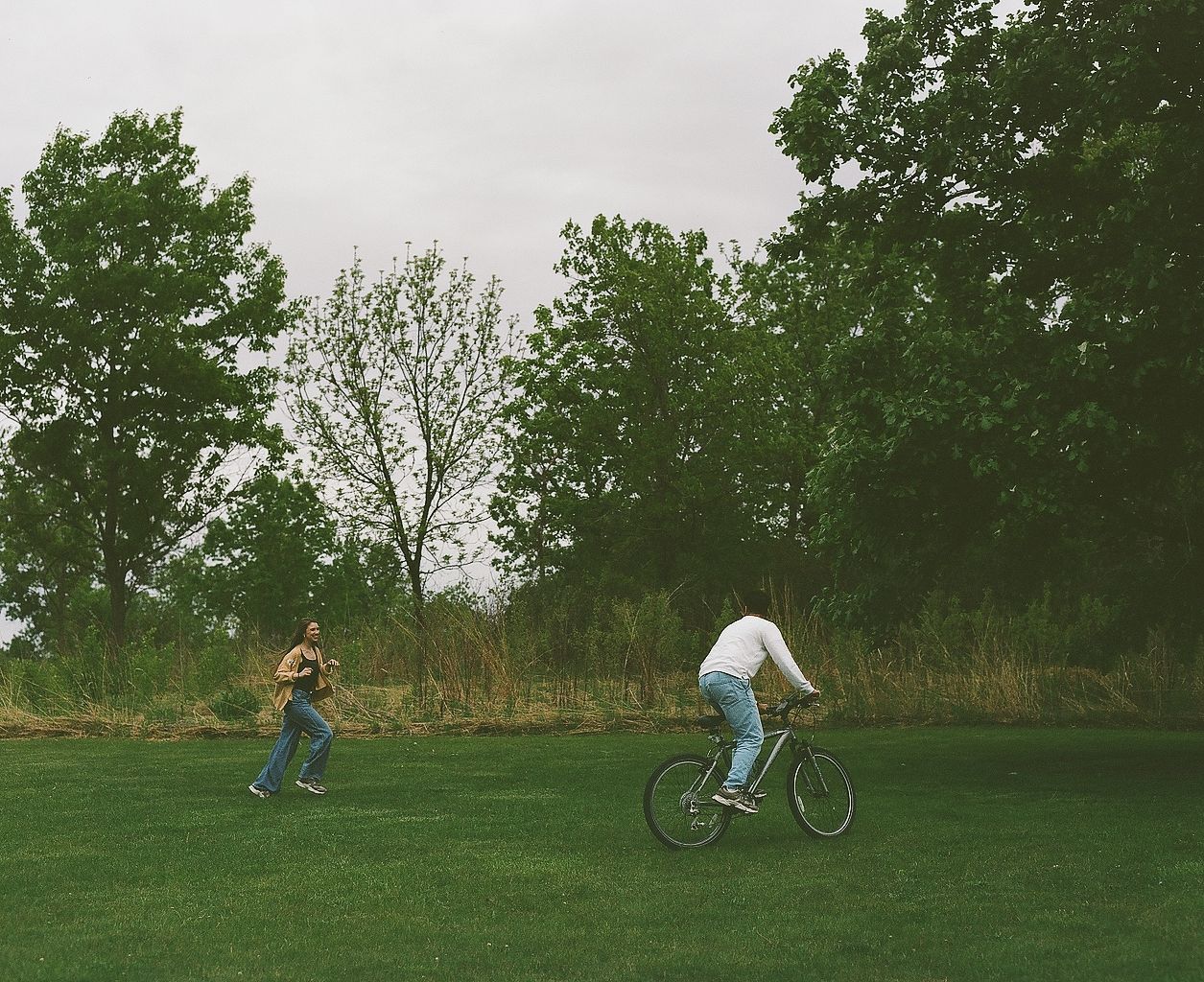 Two people enjoying a sunny day in a green park, one riding a bicycle and another running among tall trees.