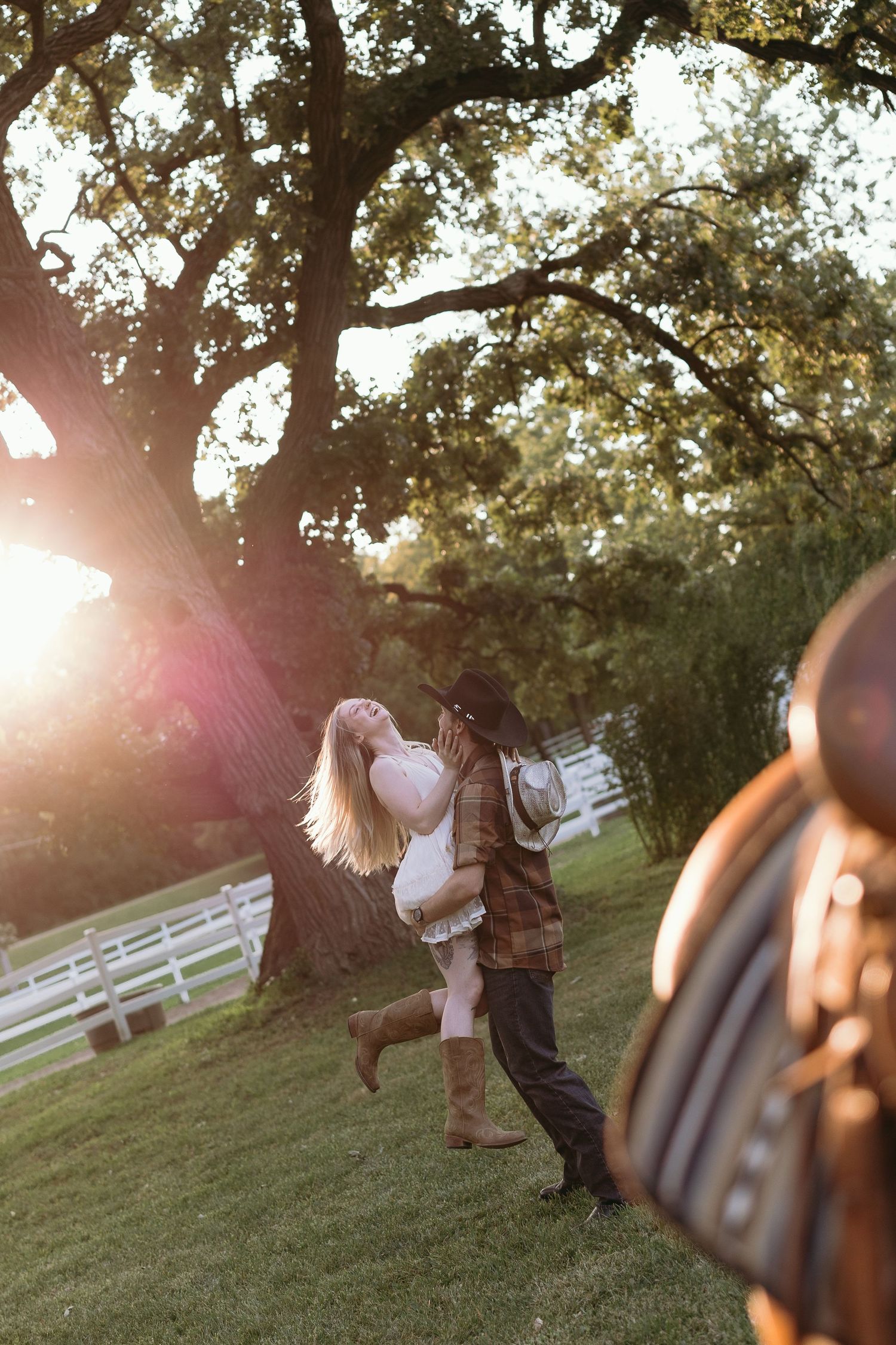 Couple dances and embraces in golden sunlit field with white fence and trees in background.