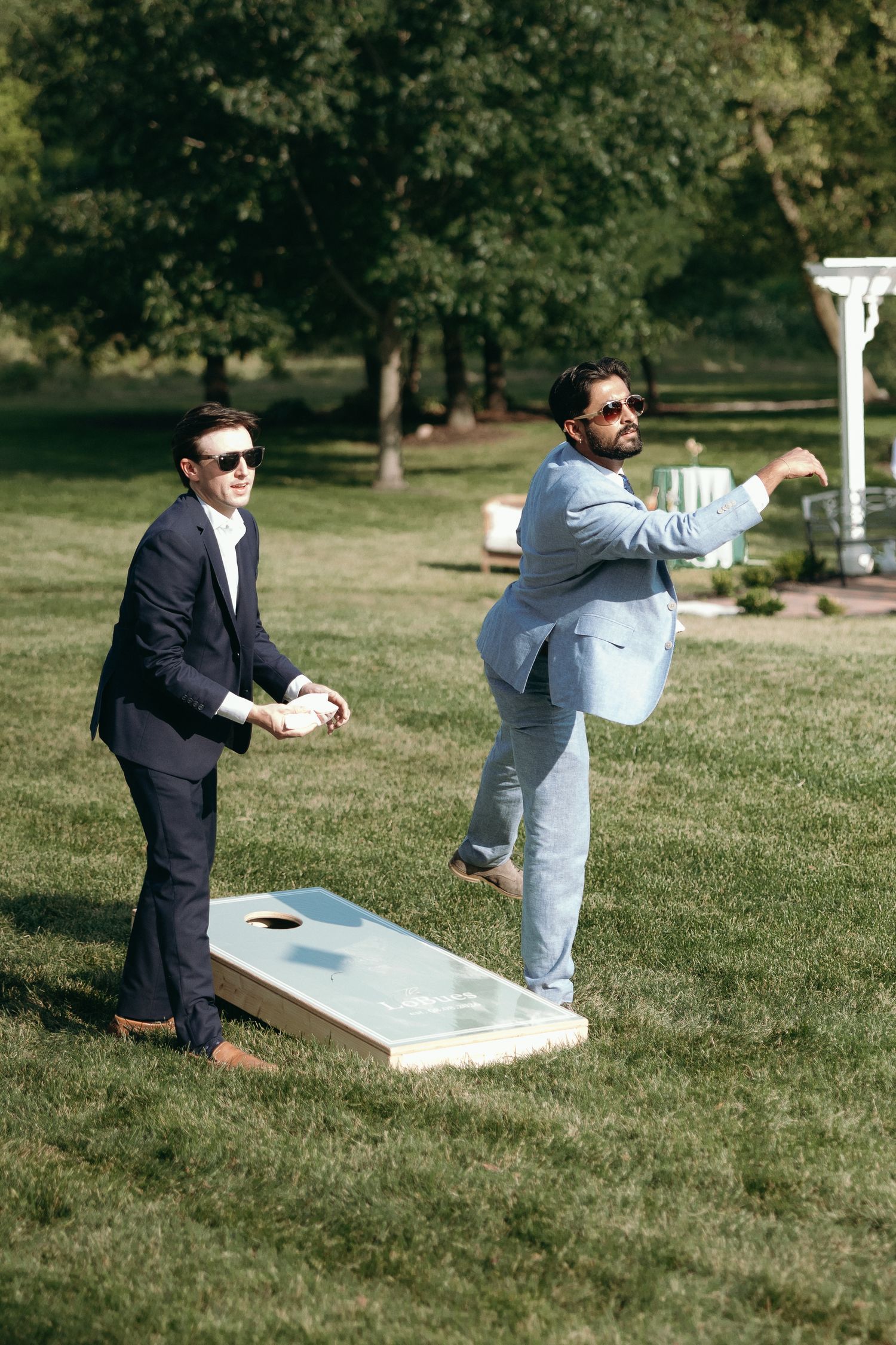 Two men in suits play cornhole lawn game at outdoor wedding reception on sunny day.