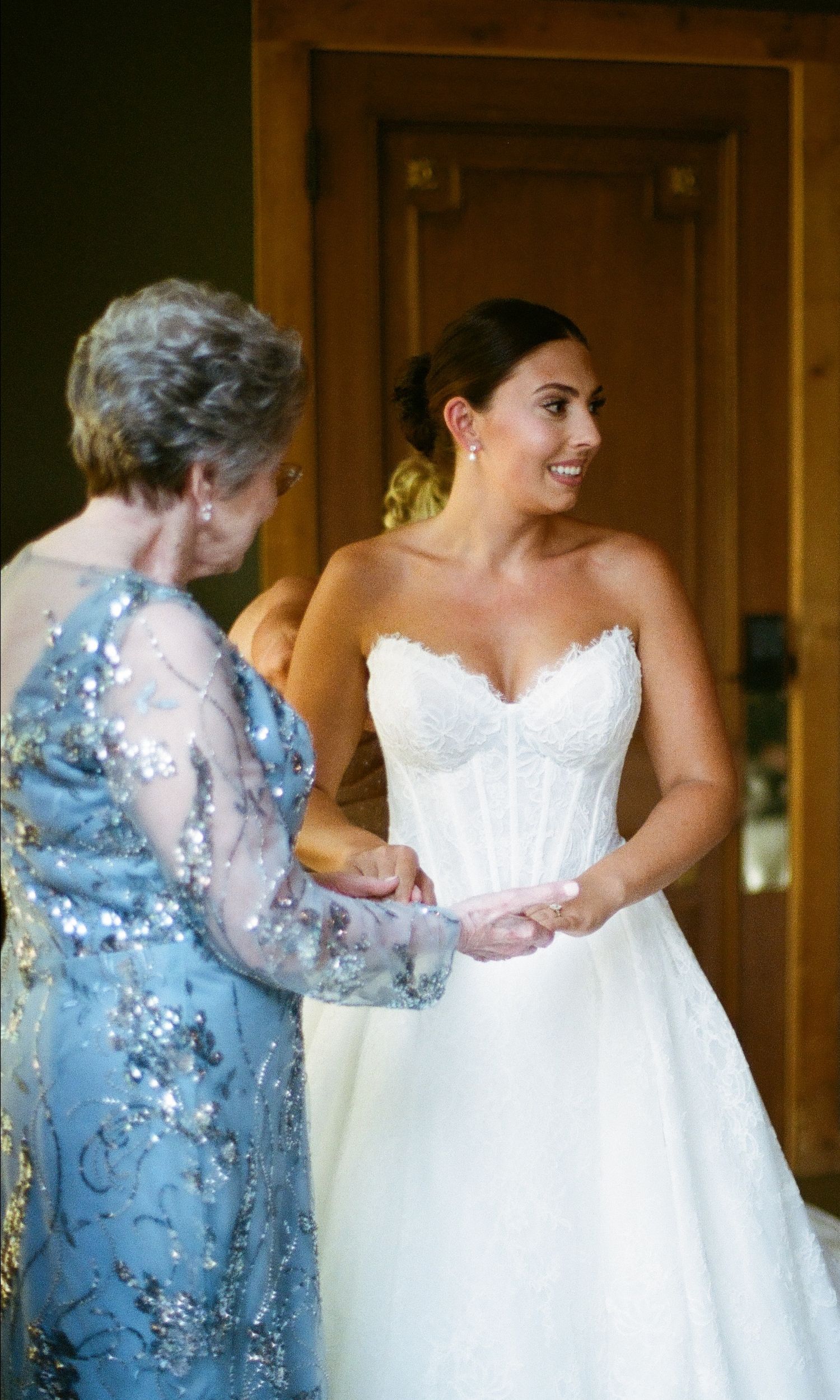 Bride in white strapless lace wedding gown smiling while elderly woman in blue sequined dress holds her hands.