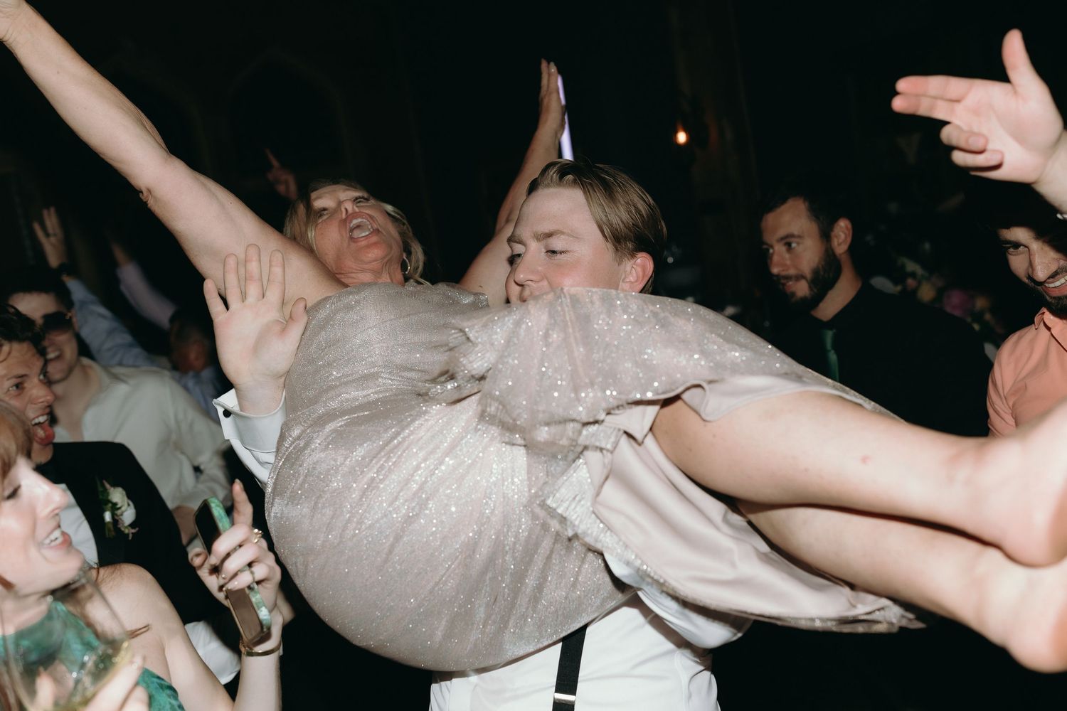 Groom lifting bride in silver dress above the crowd on a packed wedding reception dance floor.