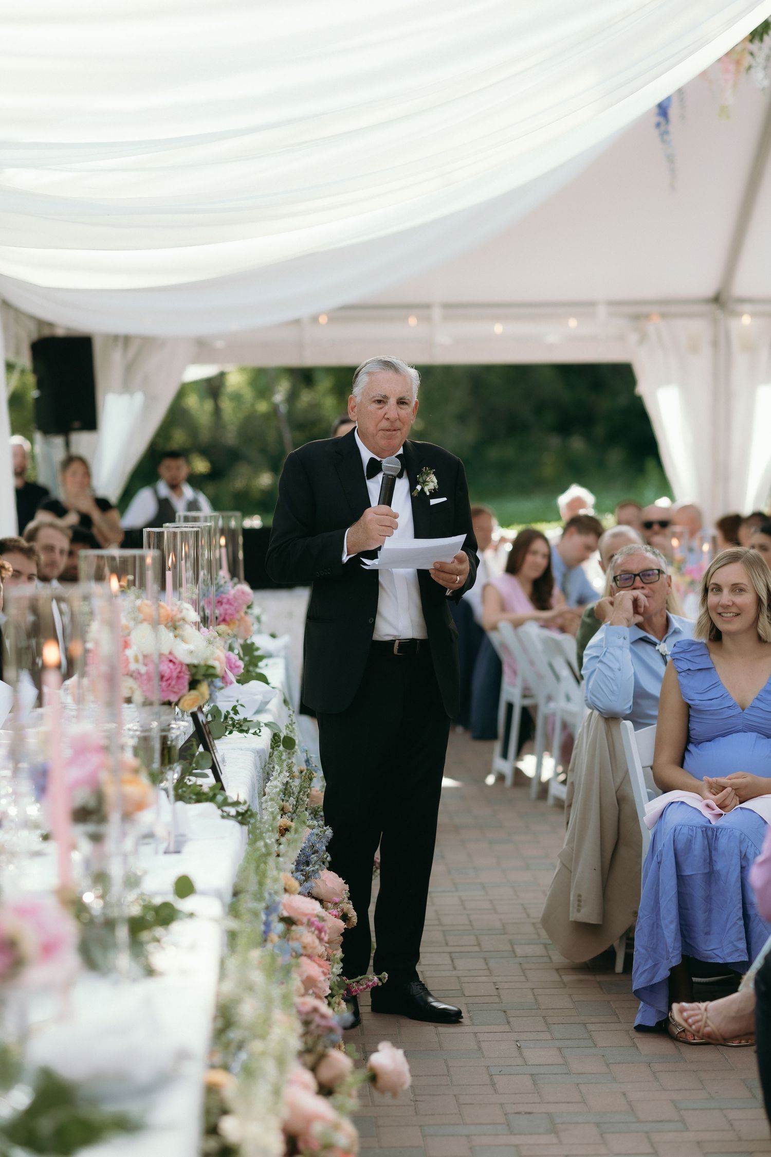 Elegant man in tuxedo giving wedding speech at floral-decorated reception table under white tent.