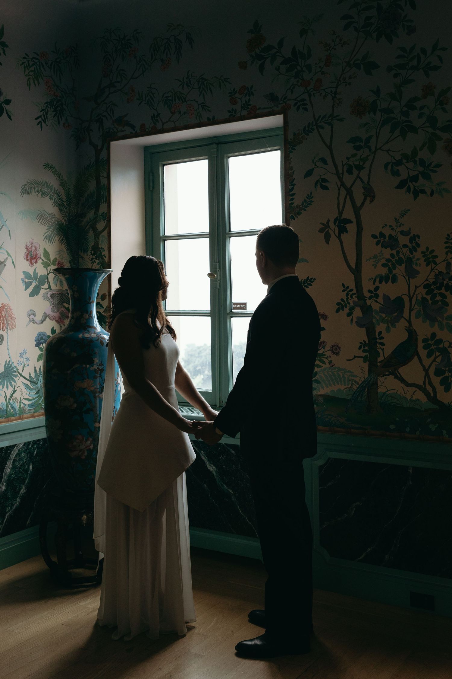 Figures in silhouette stand in dark room with decorative floral wallpaper and windows casting dramatic lighting.