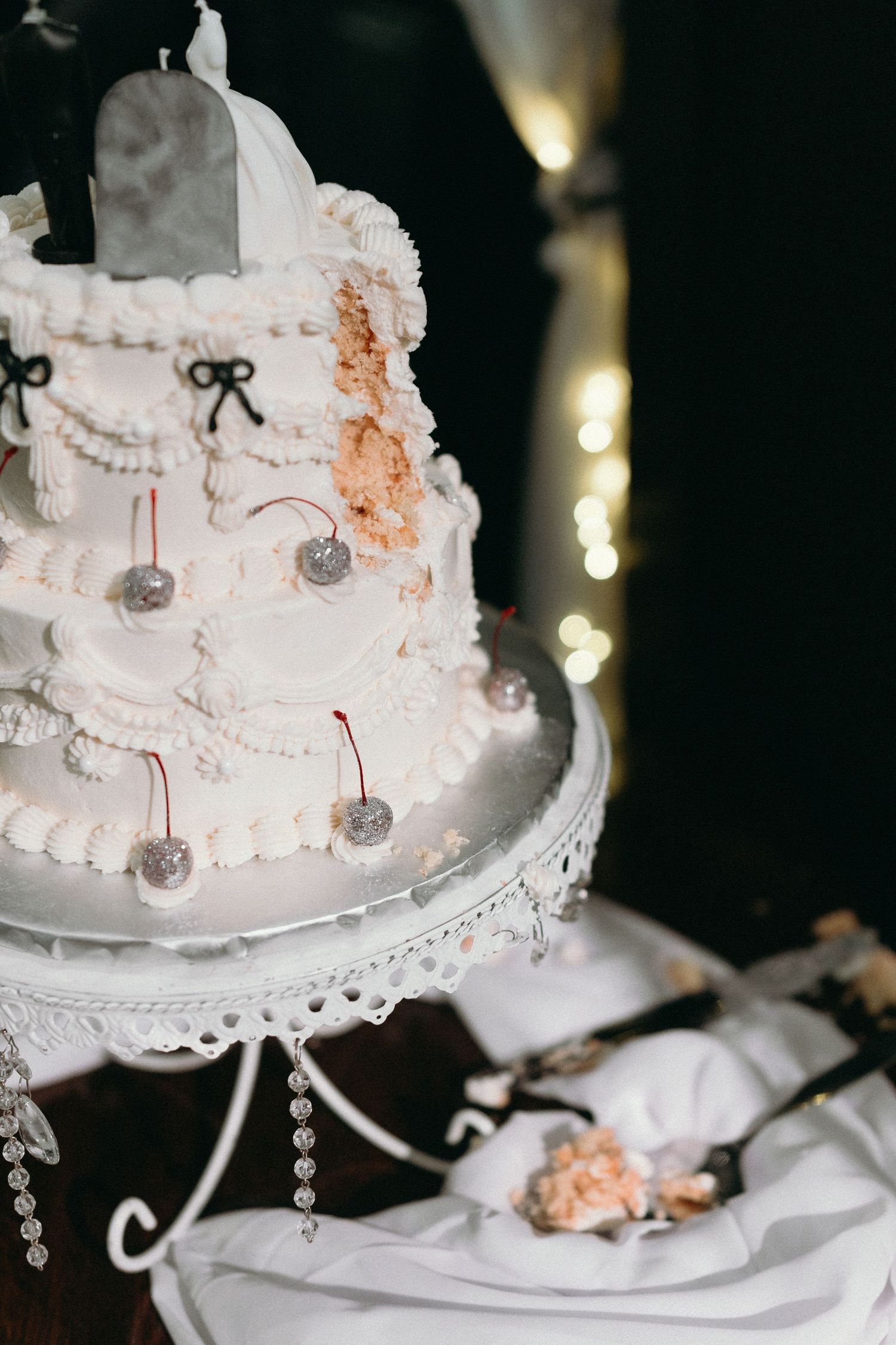 White tiered wedding cake decorated with silver cherries and delicate bows on an ornate crystal stand.