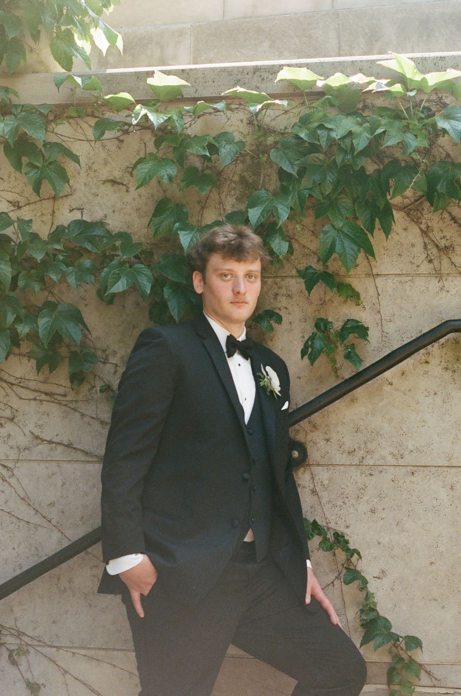 Young man in black tuxedo with boutonniere poses on stairs beside ivy-covered stone wall.