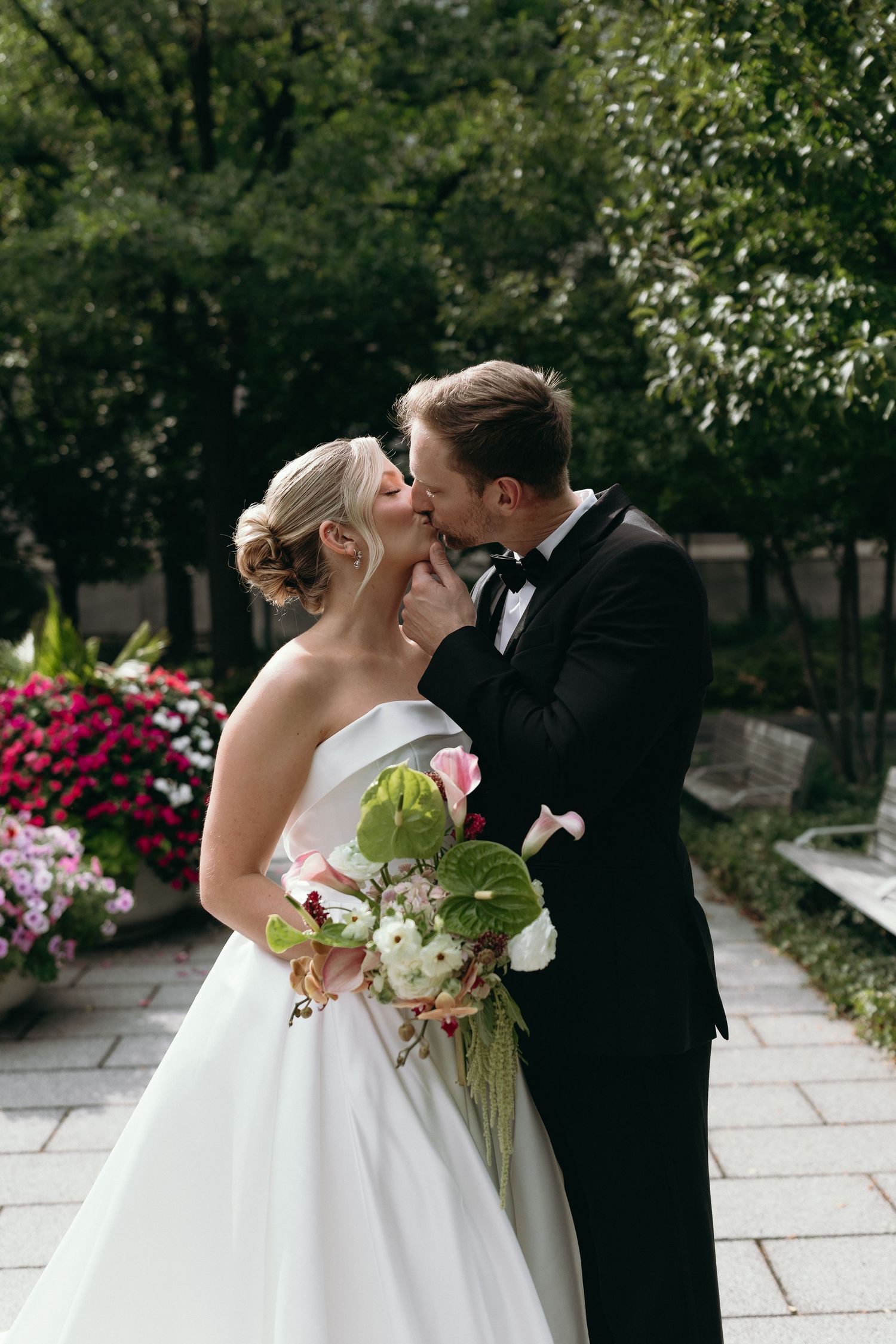 A romantic wedding moment captured in a garden setting with pink flowers and greenery as the couple shares a tender kiss.