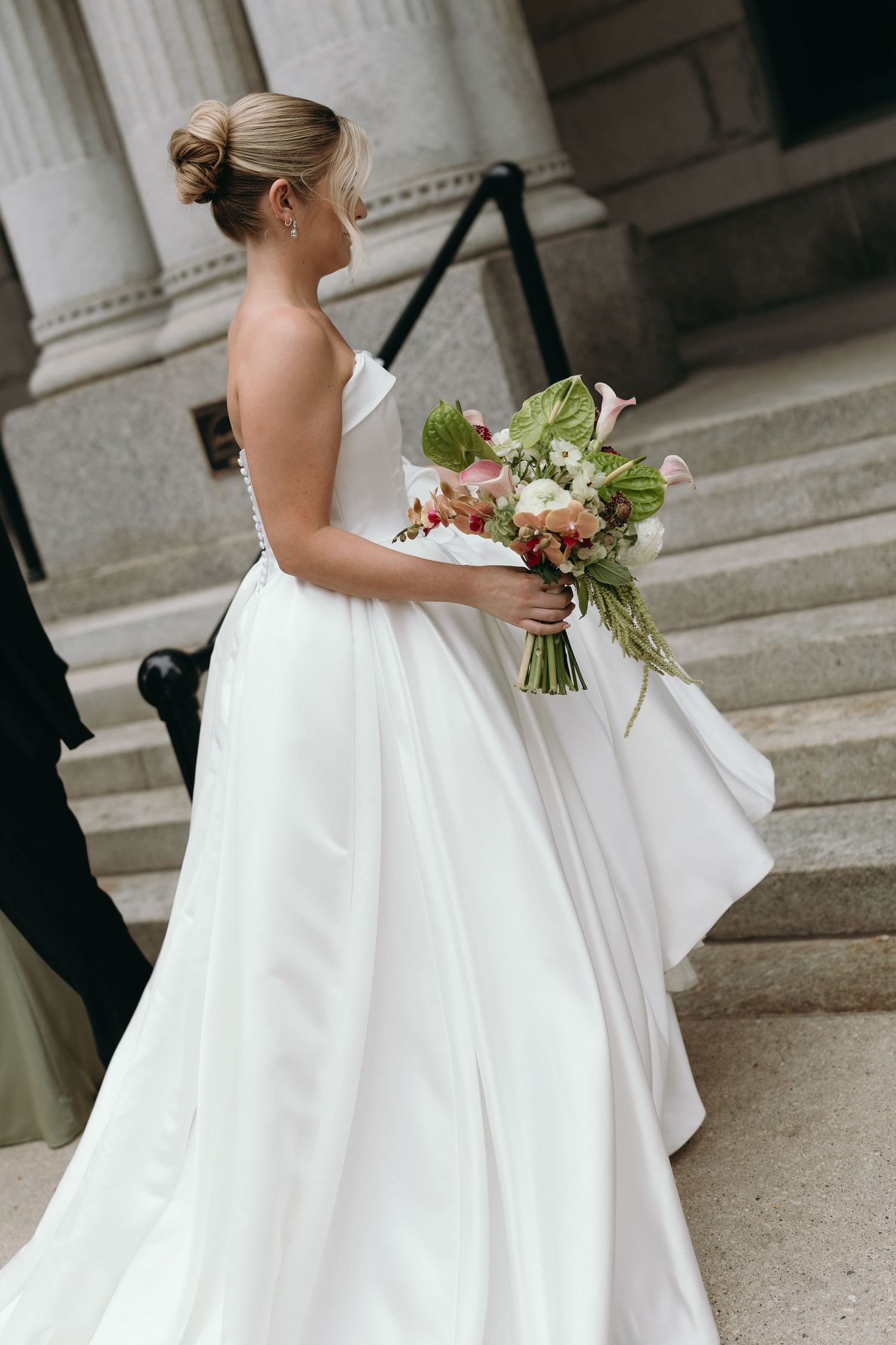 A bride in a flowing white strapless gown holds a beautiful bouquet of flowers while standing on stone steps.