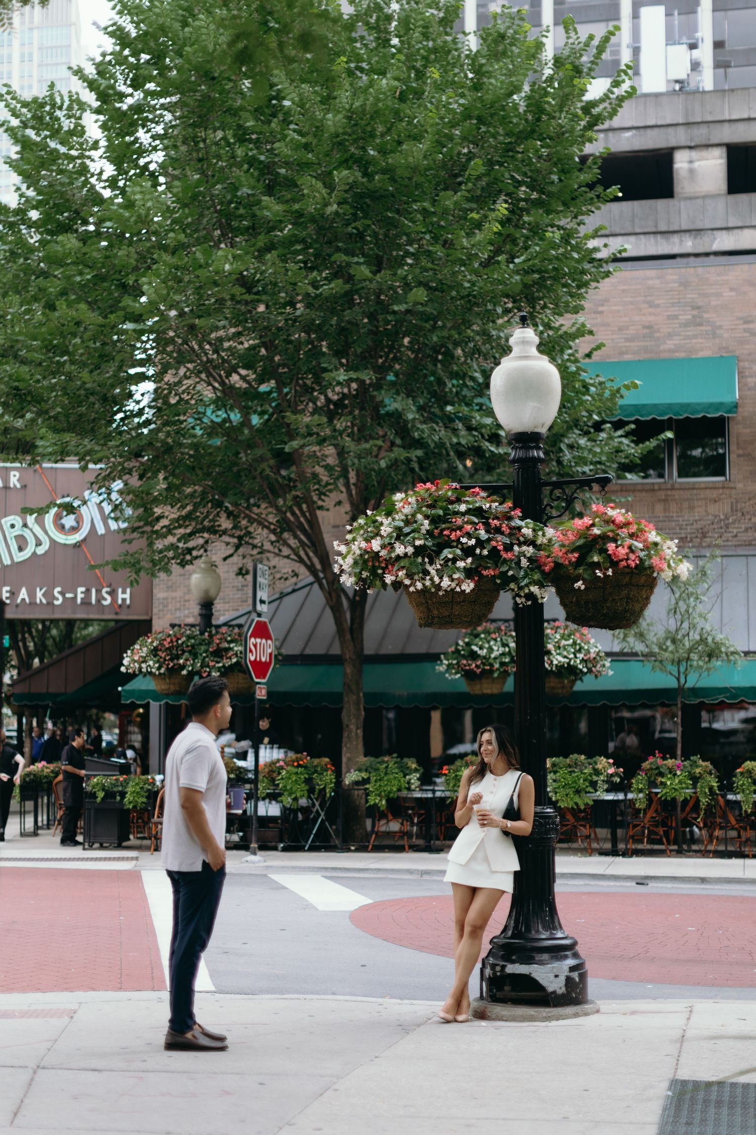 People gather and walk along a brick-paved city plaza with trees and retail storefronts in the background.