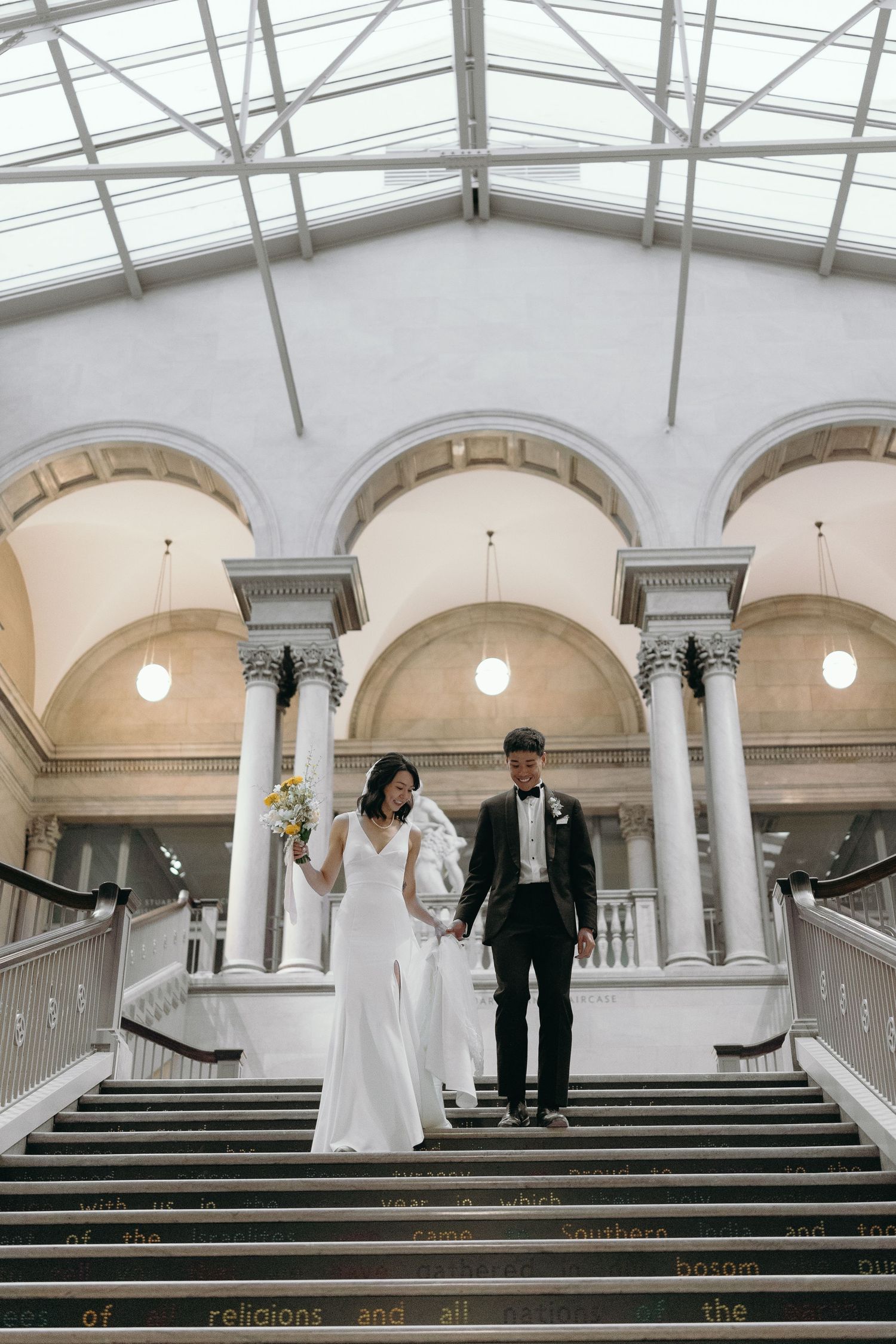 A couple standing on grand marble steps beneath an arched ceiling with columns.