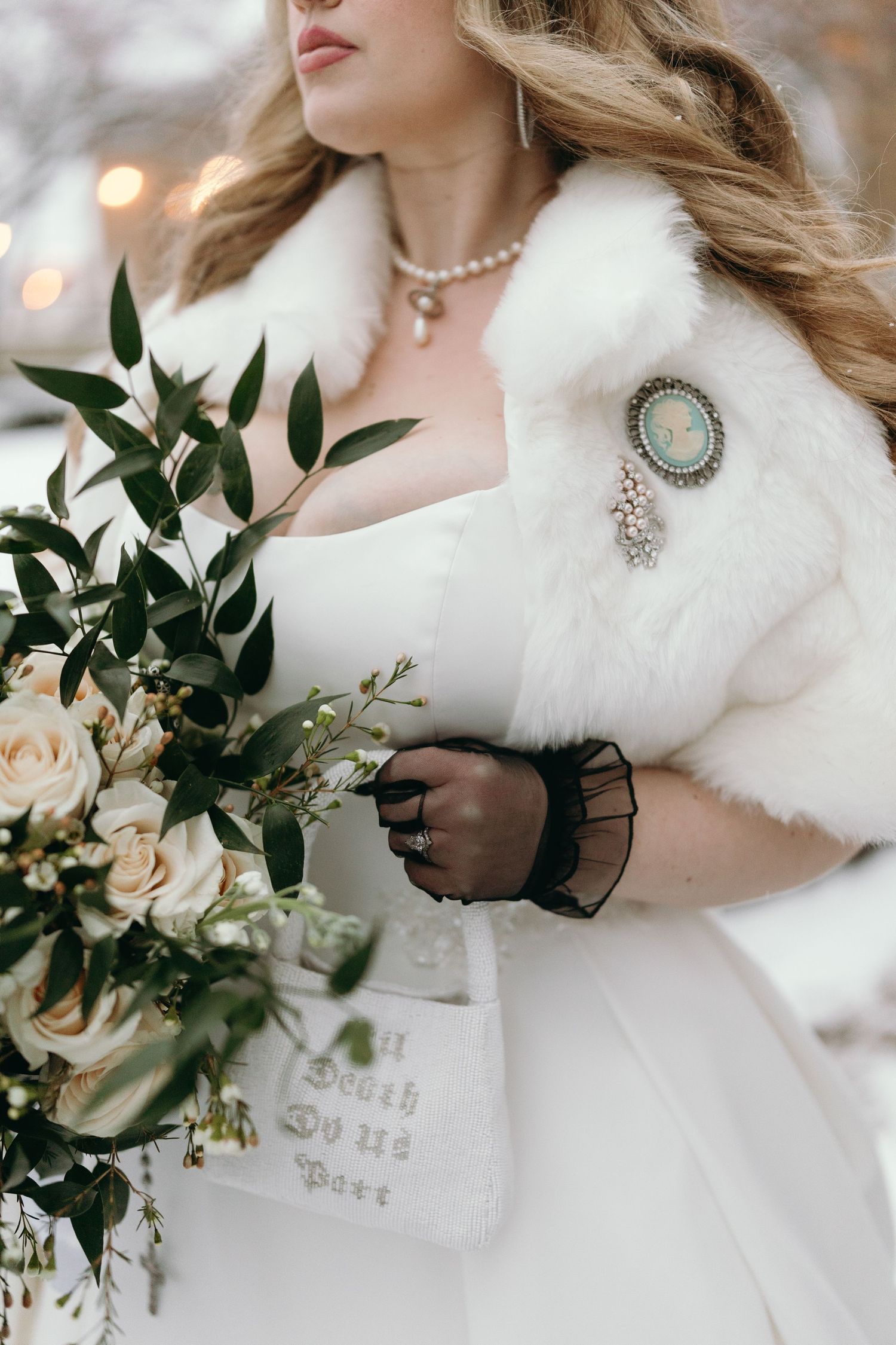 Wedding bouquet with cream roses and greenery held against white fur wrap with vintage brooch.