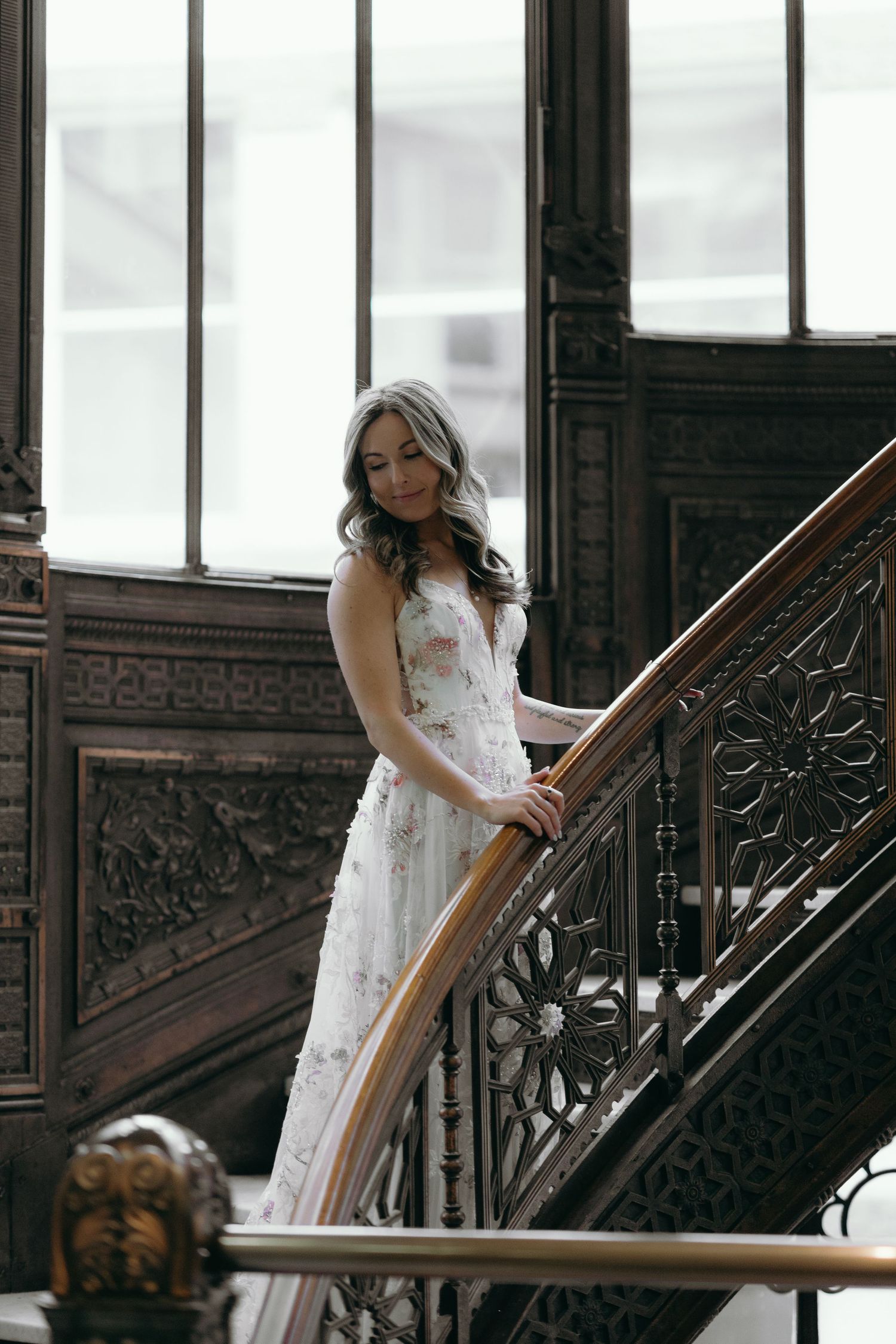 Ornate dark wood staircase in a historic building with large windows providing natural light.