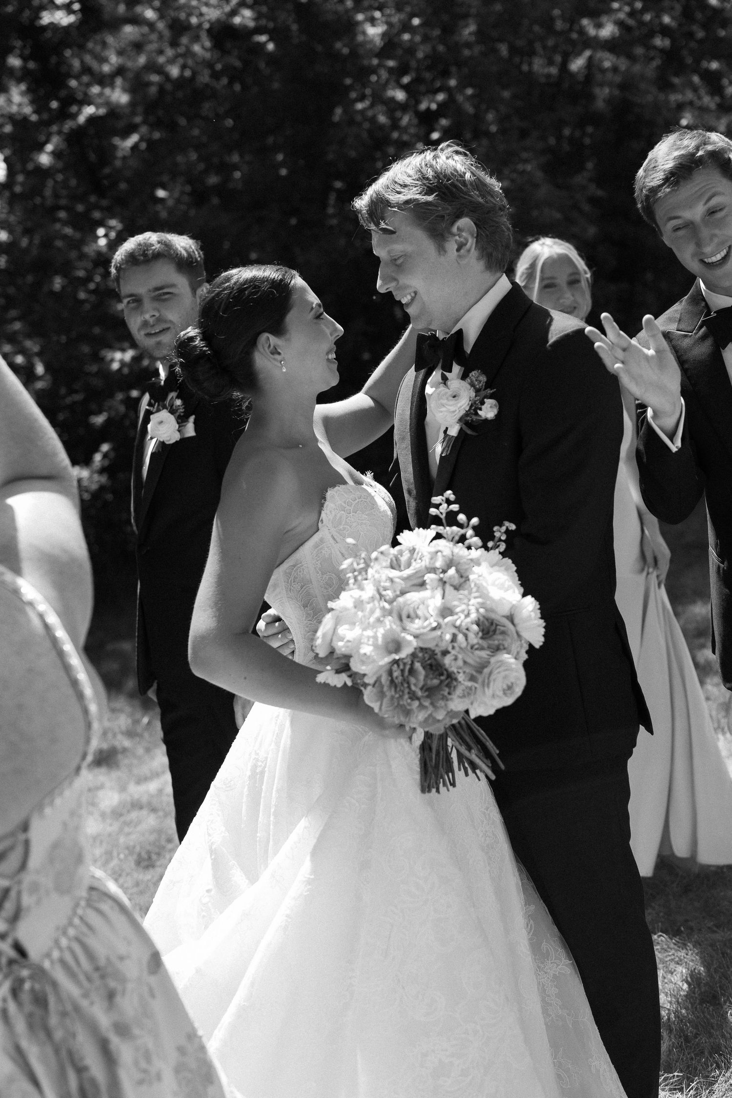 Black and white photo of smiling bride and groom sharing a joyful moment surrounded by wedding party outdoors.