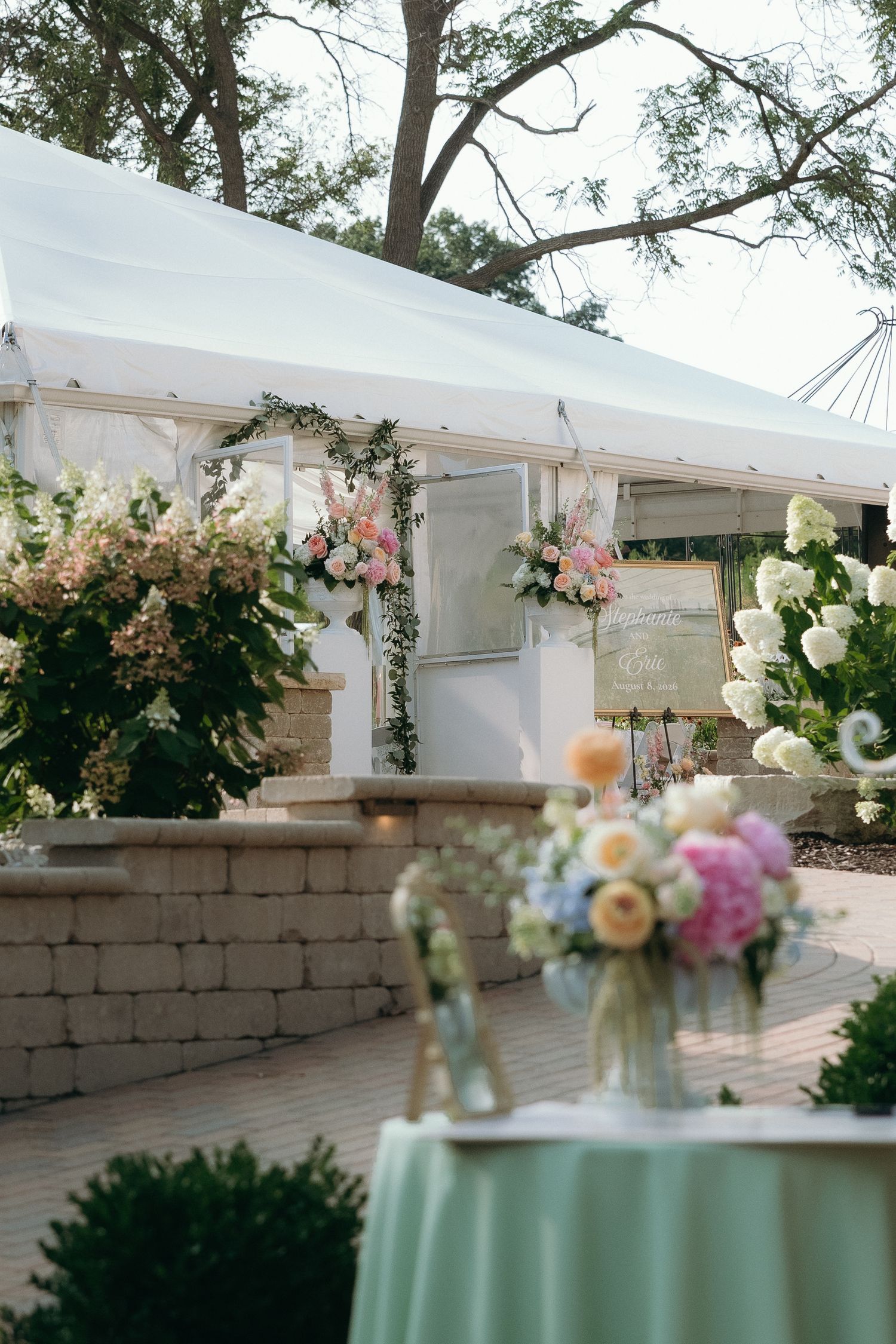 Elegant outdoor wedding tent venue with floral arrangements, pastel blooms, and a mint green cocktail table.