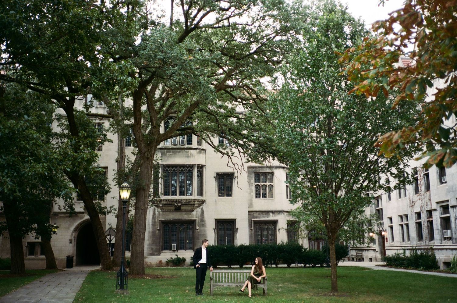 Two people relaxing on a bench in a lush green courtyard beside a classic stone university building.