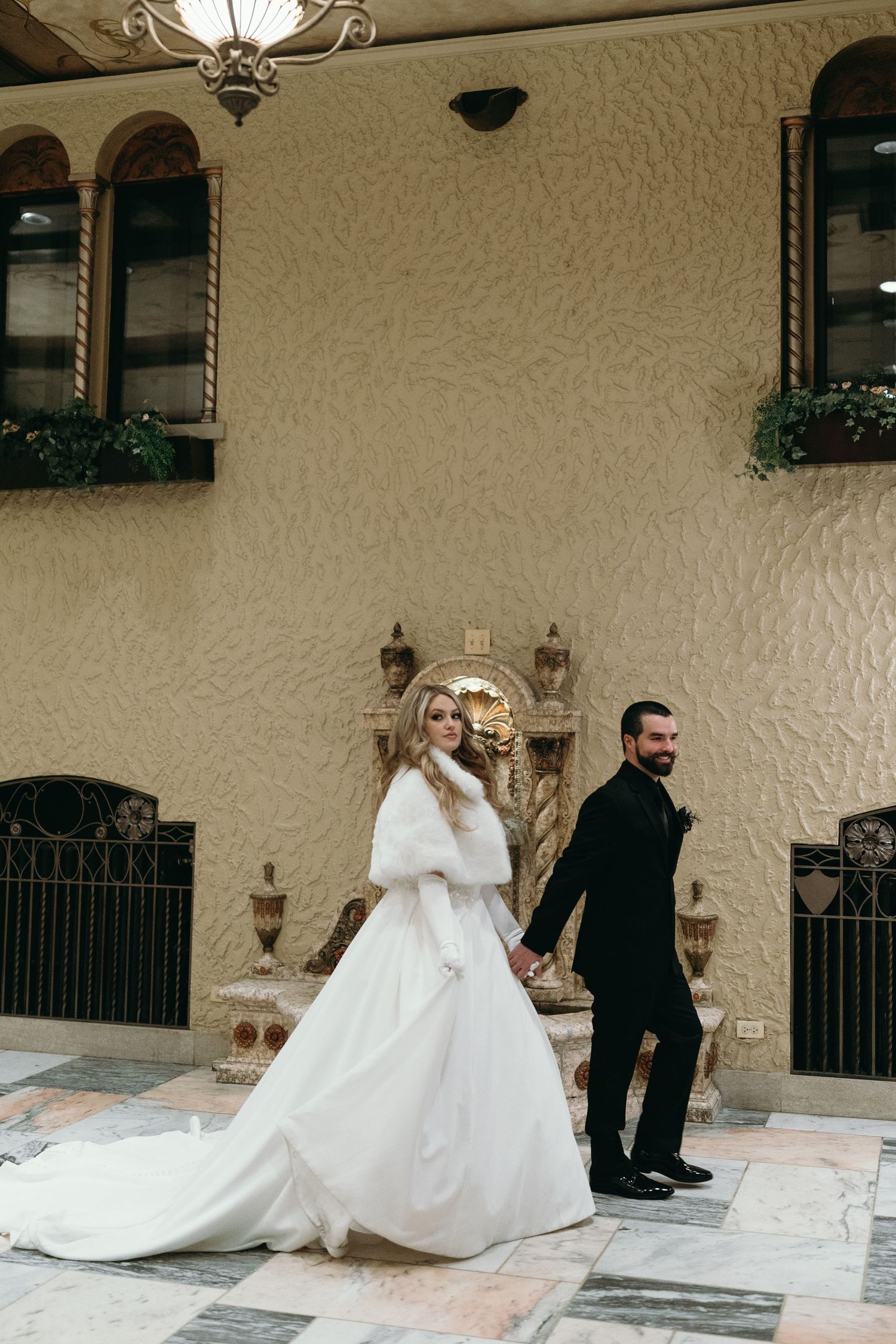 A couple in wedding attire walk hand in hand through an elegant archway with Mediterranean style decor.