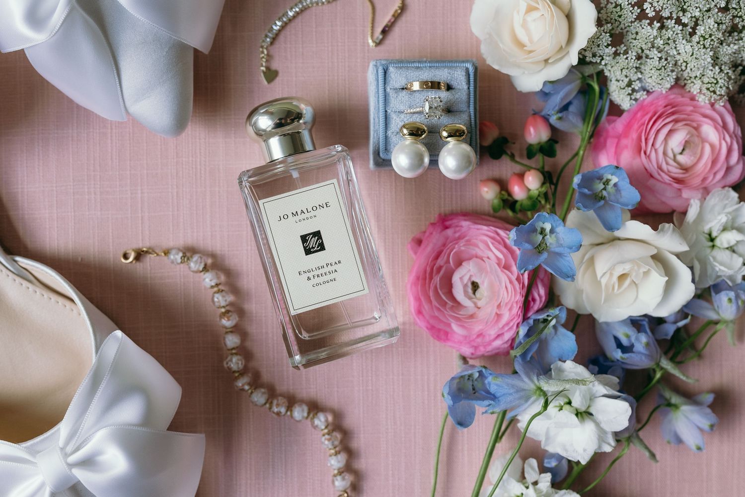 Wedding accessories including perfume bottles, pearls, and delicate flowers arranged on a pink surface.