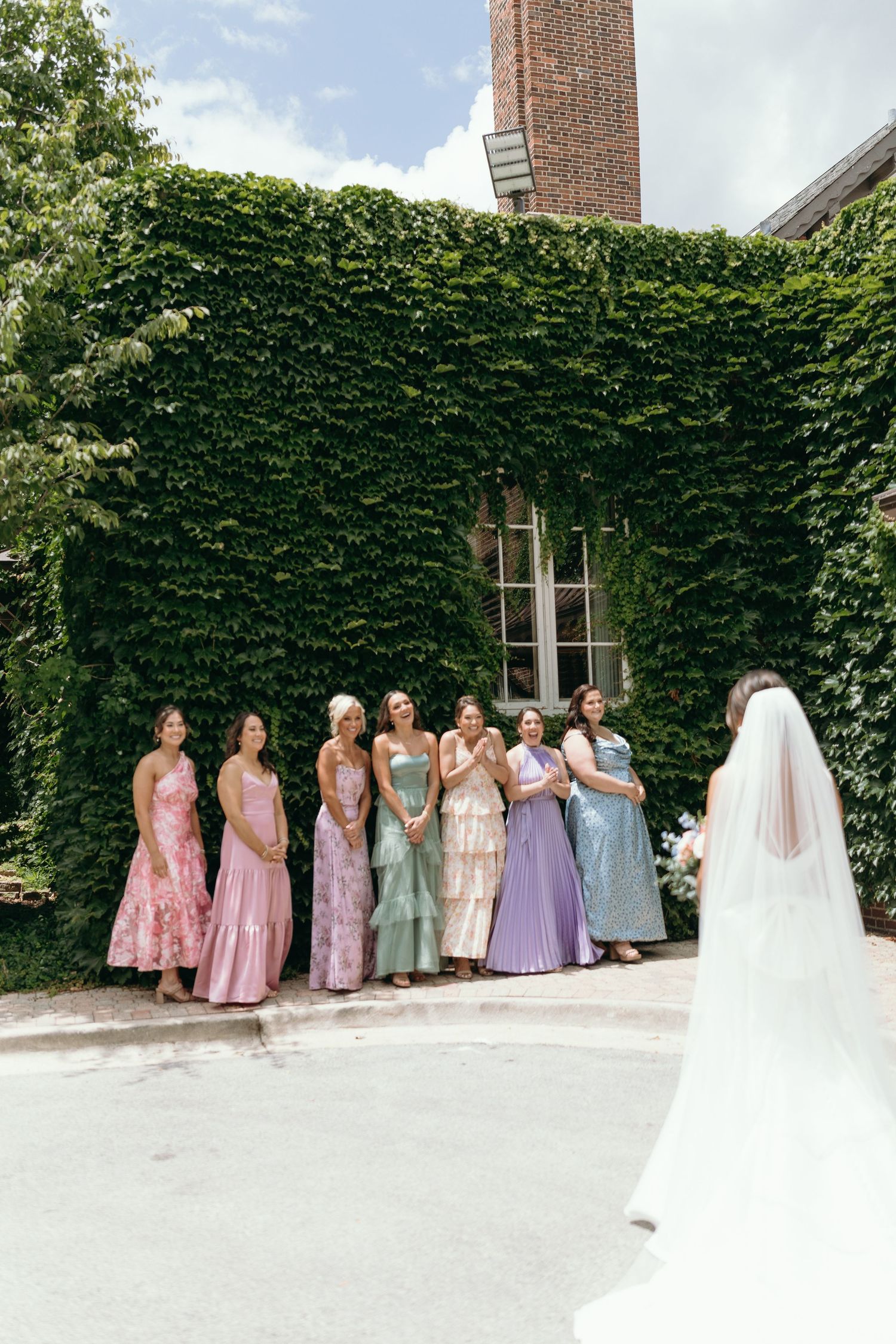 Line of bridesmaids in pastel dresses posing against ivy-covered wall with bride in white.