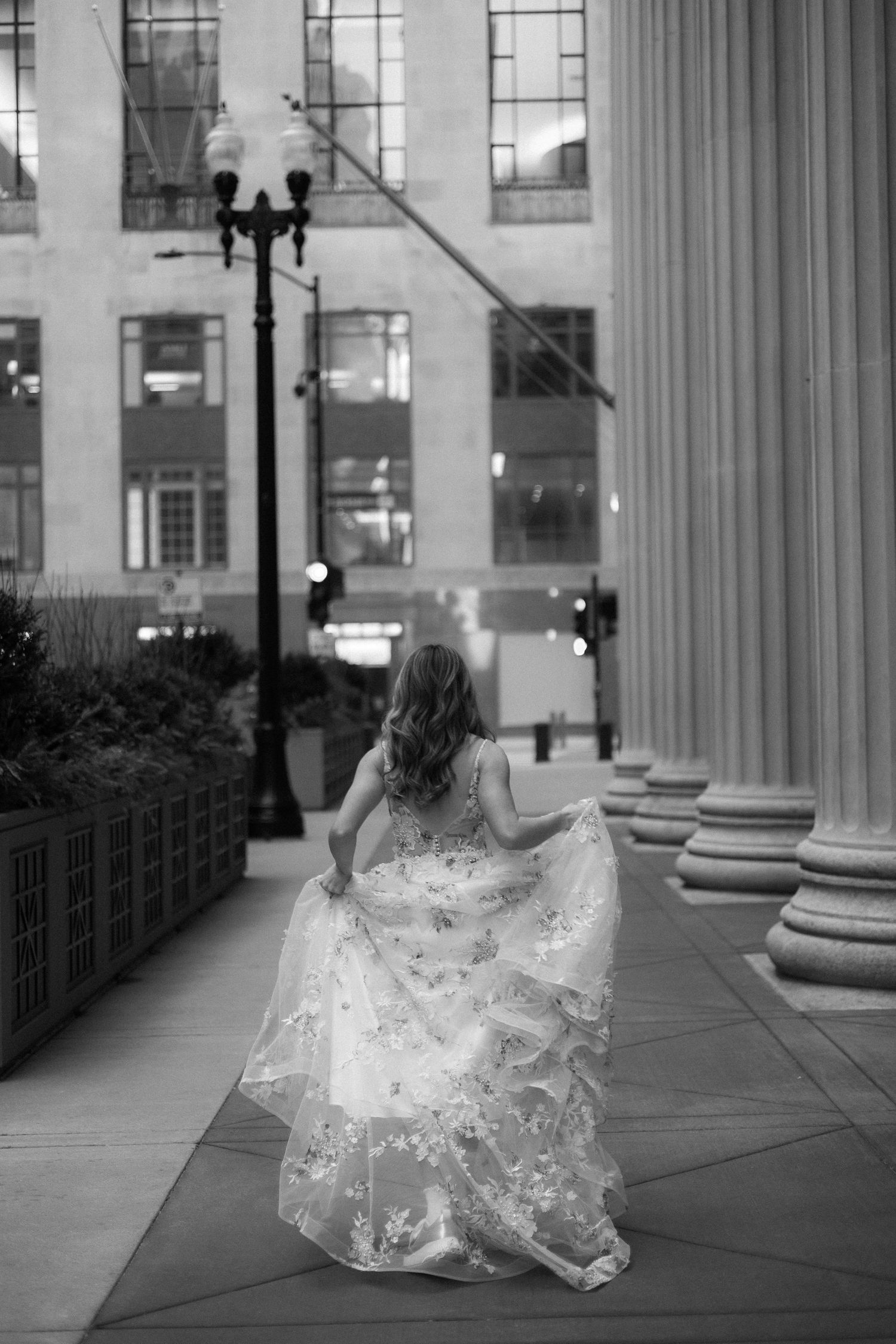 White wedding dress flowing on urban sidewalk against classical architecture with columns.