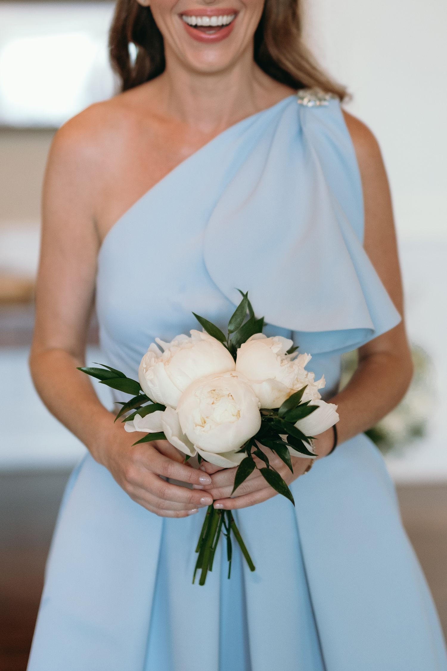 Someone wearing a light blue one-shoulder bridesmaid dress holds a simple bouquet of white peonies with greenery.