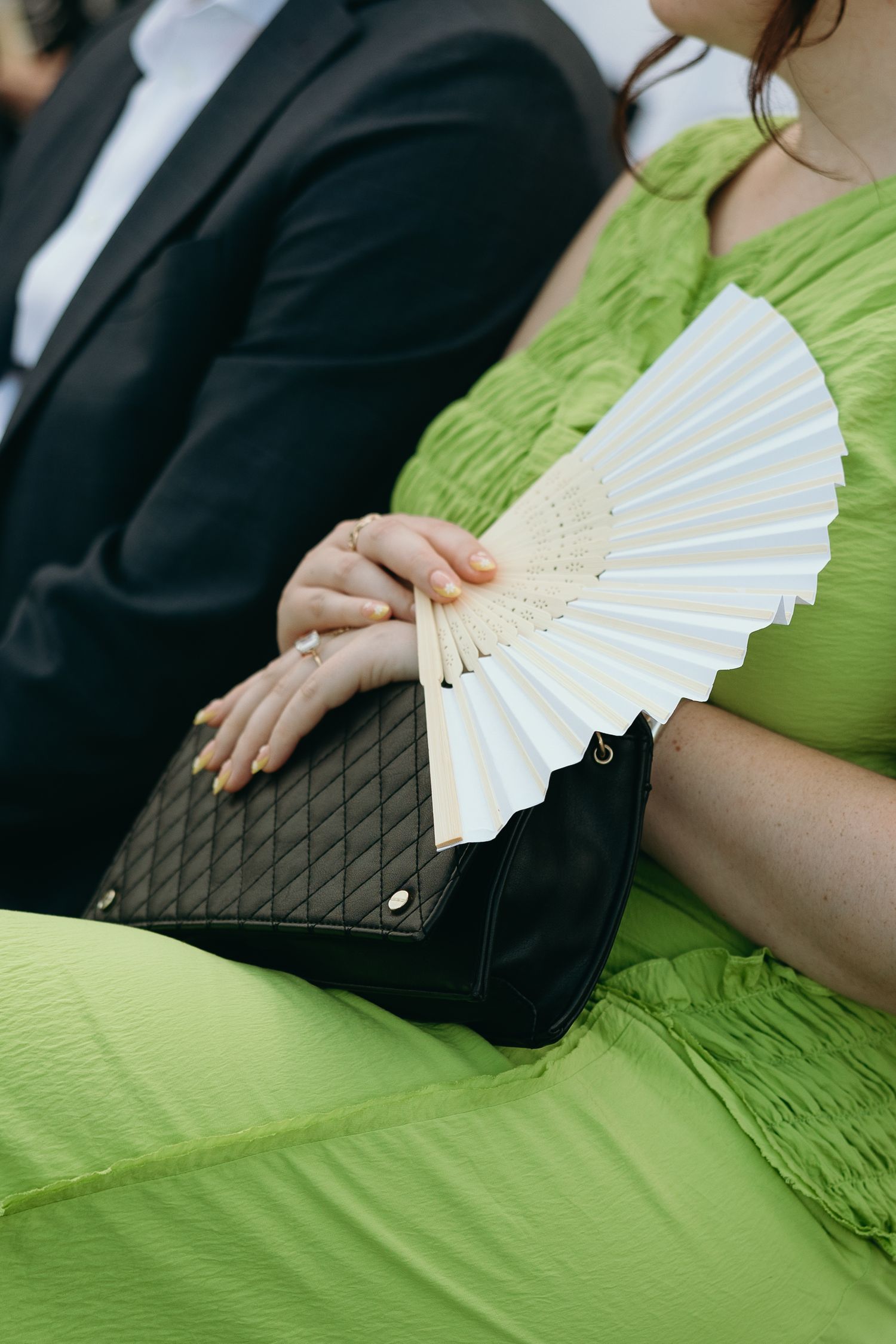 Person in lime green dress holds white folding fan while carrying black quilted handbag next to someone in black suit.