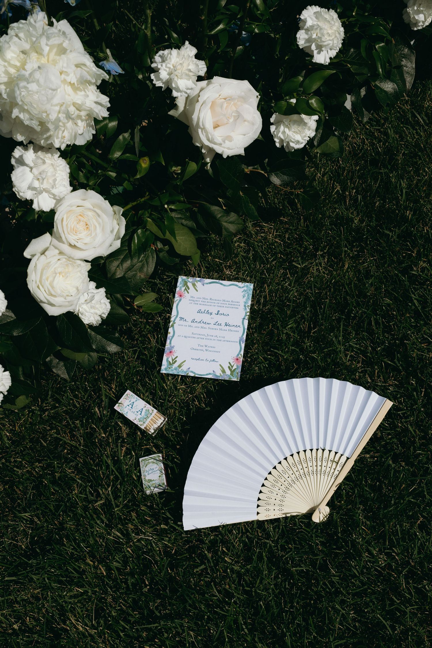 White paper invitations and folding hand fans arranged with white roses against dark foliage background.