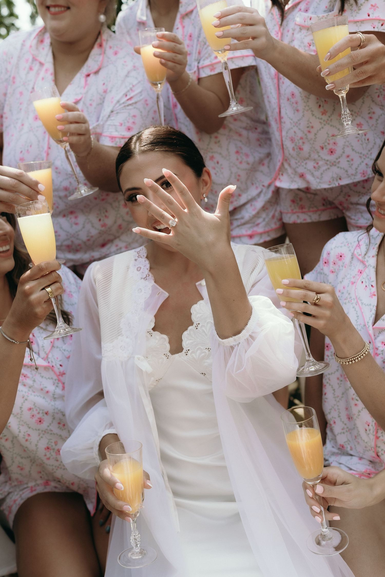 Bride in white robe shows off engagement ring while holding mimosa with bridesmaids in floral pajamas.