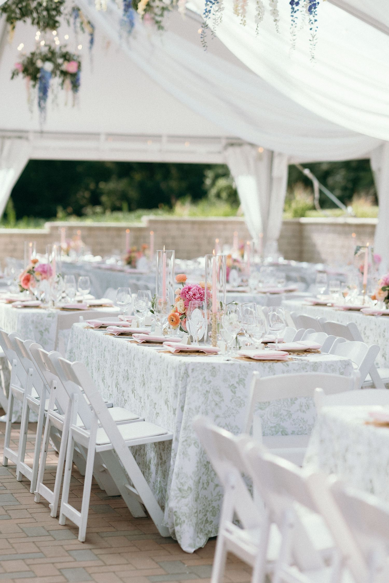 Elegant outdoor wedding reception under white tent with floral tablecloths, colorful centerpieces, and white folding chairs.