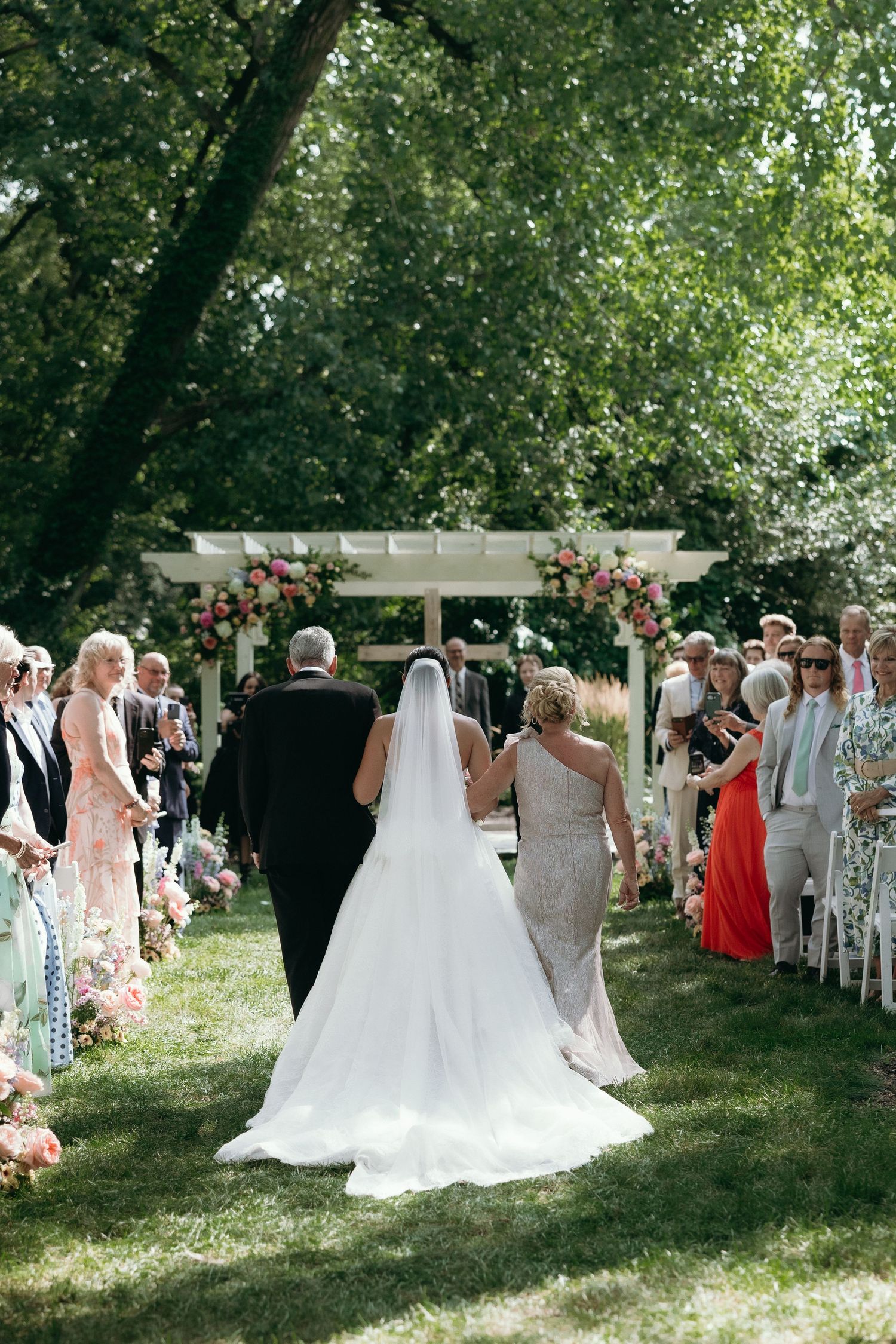 Bride walks down outdoor aisle with parents toward floral arch at garden wedding ceremony.