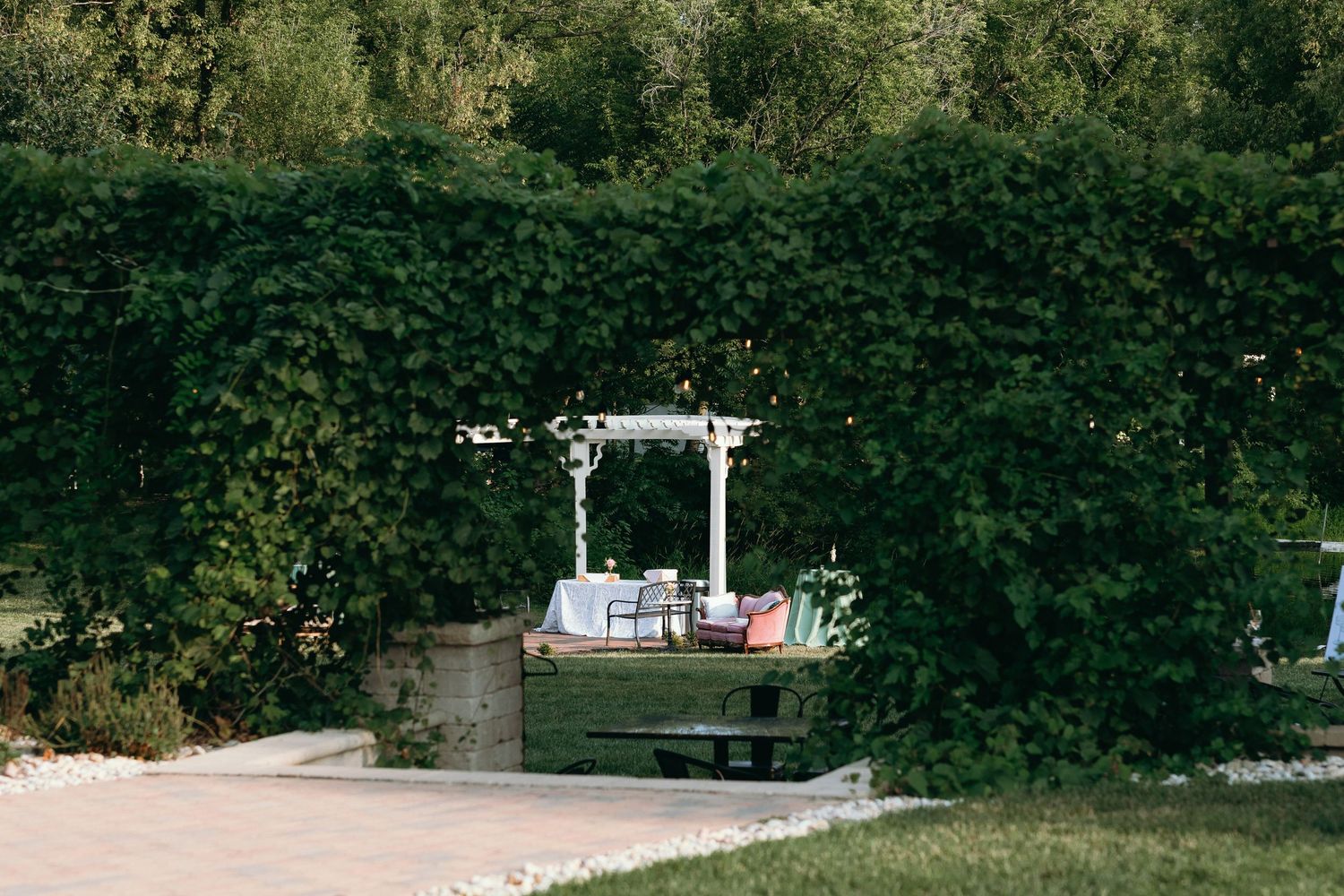 A white pergola surrounded by lush green ivy hedges with outdoor seating in a serene garden setting.