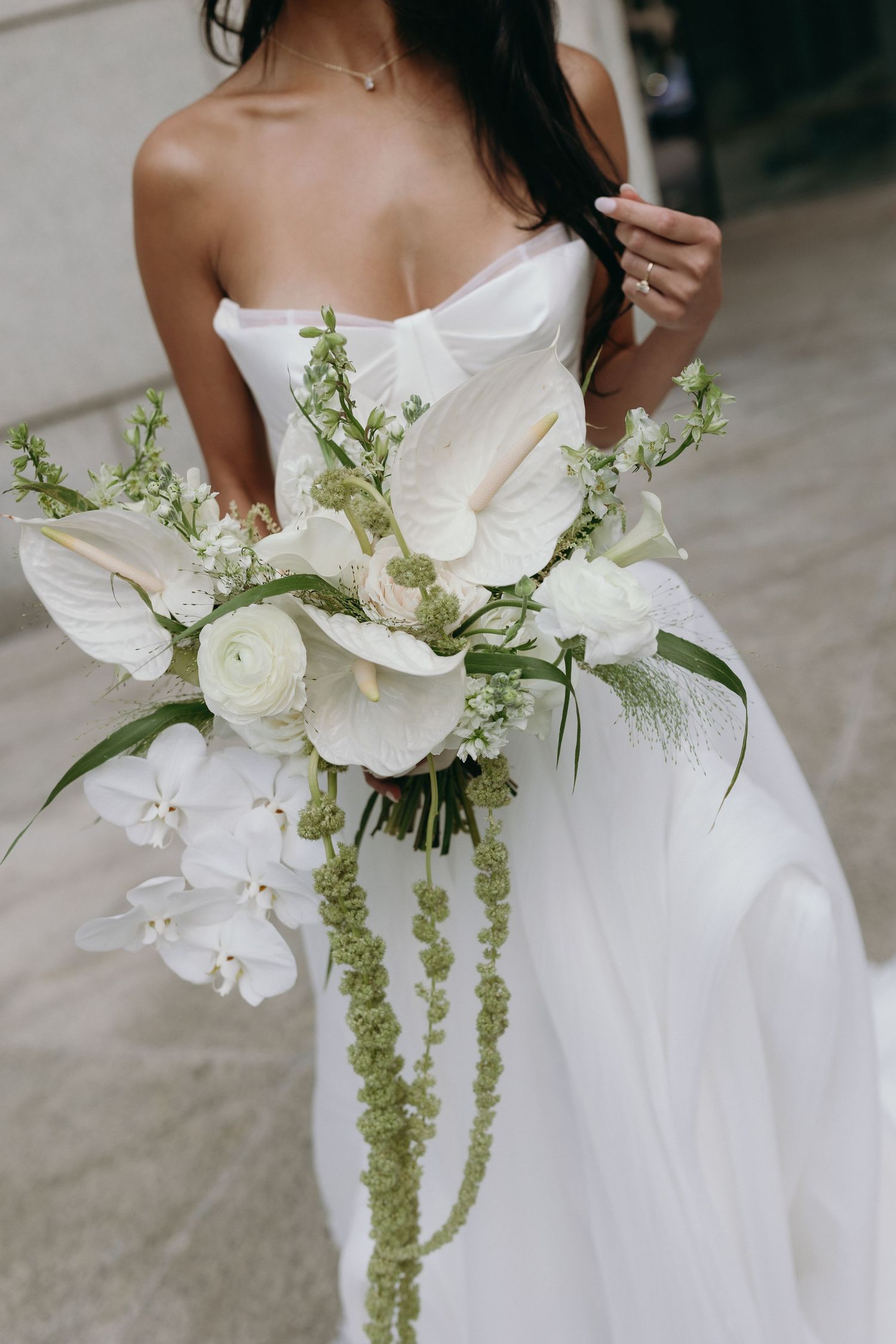 Close-up of elegant white bridal bouquet with roses, orchids and cascading green foliage against white wedding dress.
