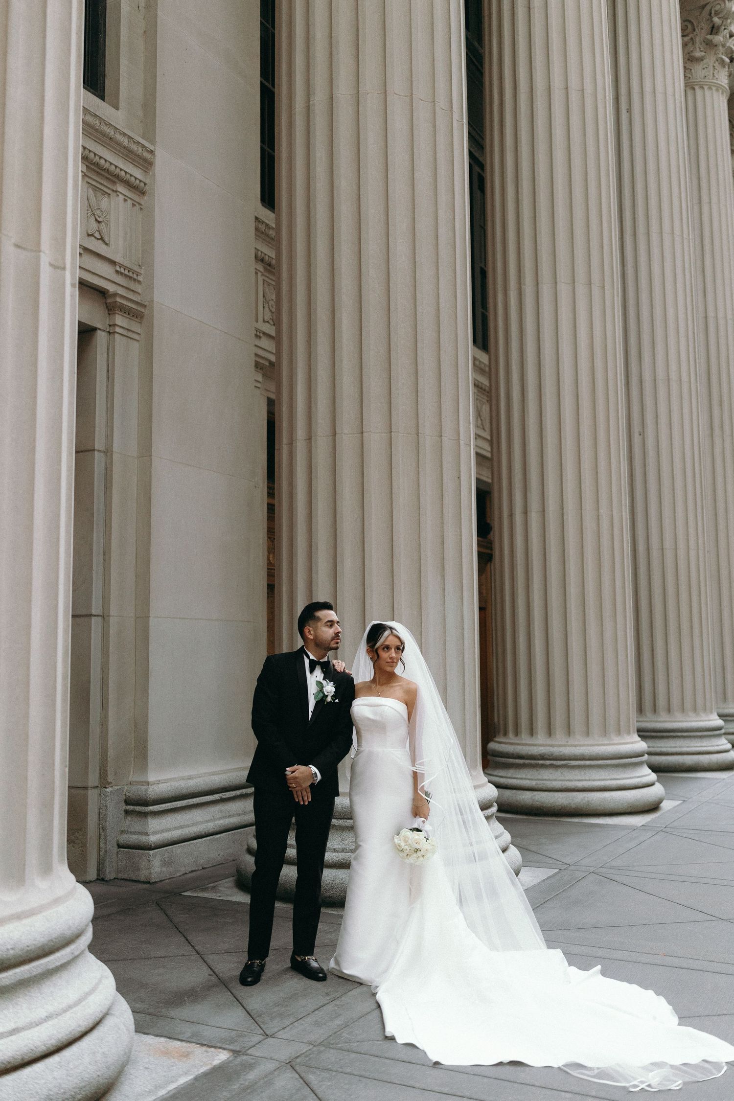 Wedding couple poses elegantly between classical white stone columns in an architectural setting.