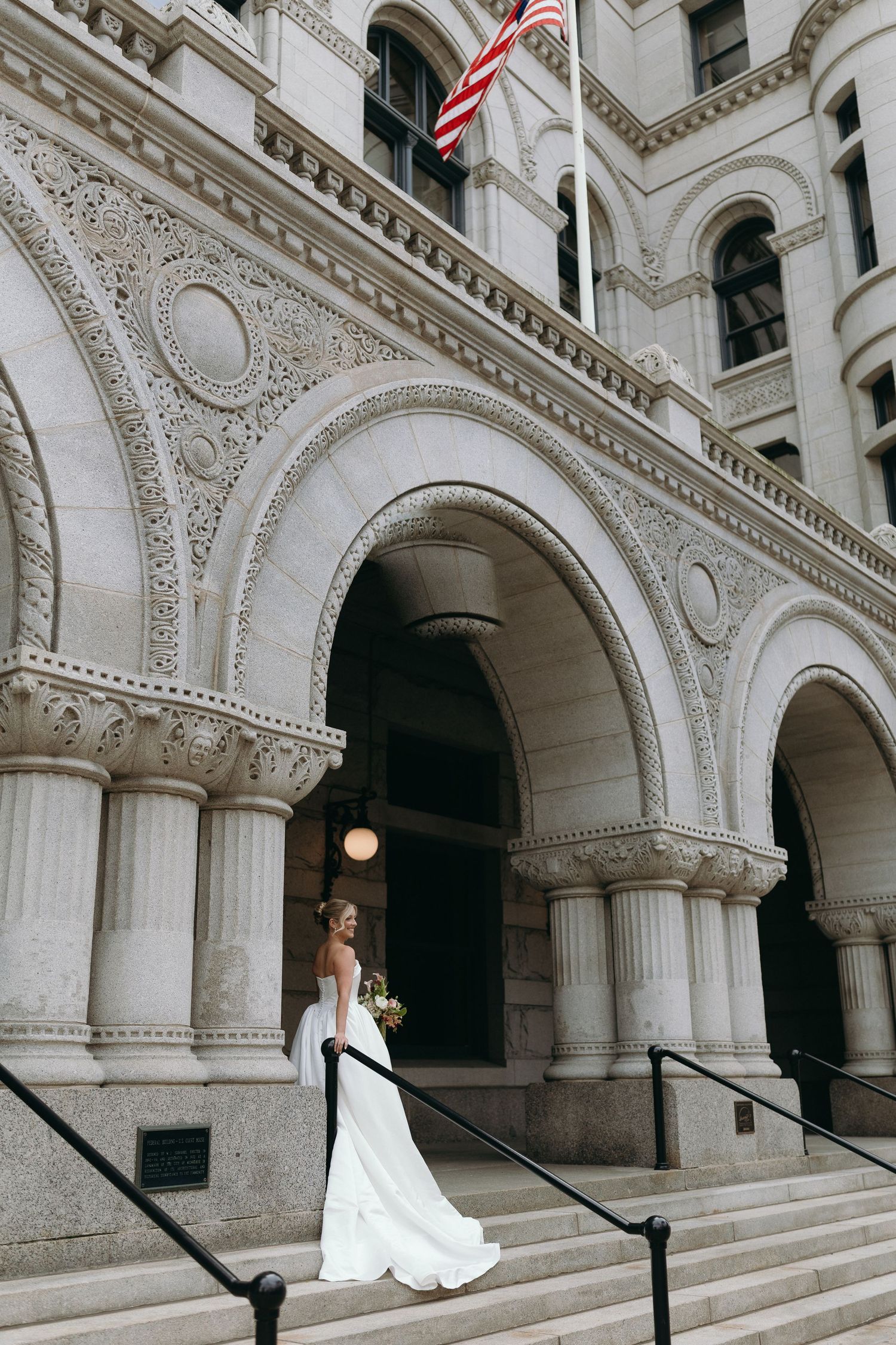 A bride in a white wedding dress poses on ornate marble steps of a historic building with elegant arched architecture.