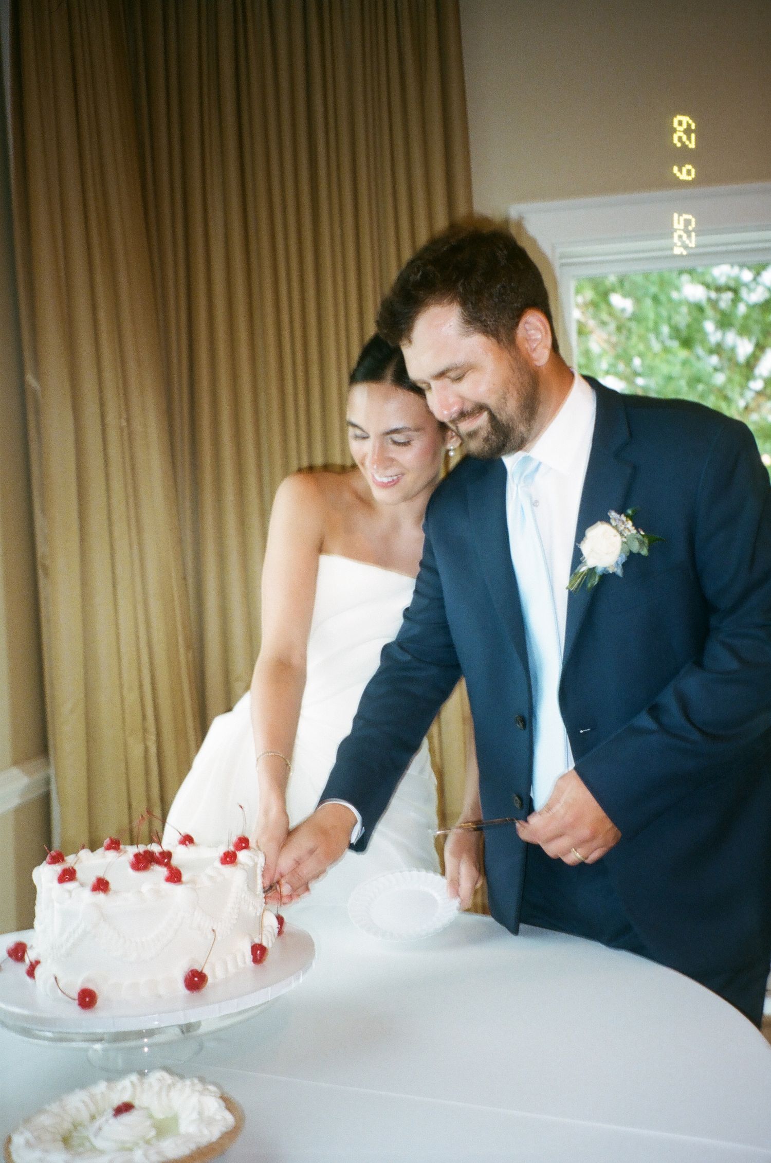 Bride and groom cutting white wedding cake with red cherries, man in navy suit, woman in strapless white gown.