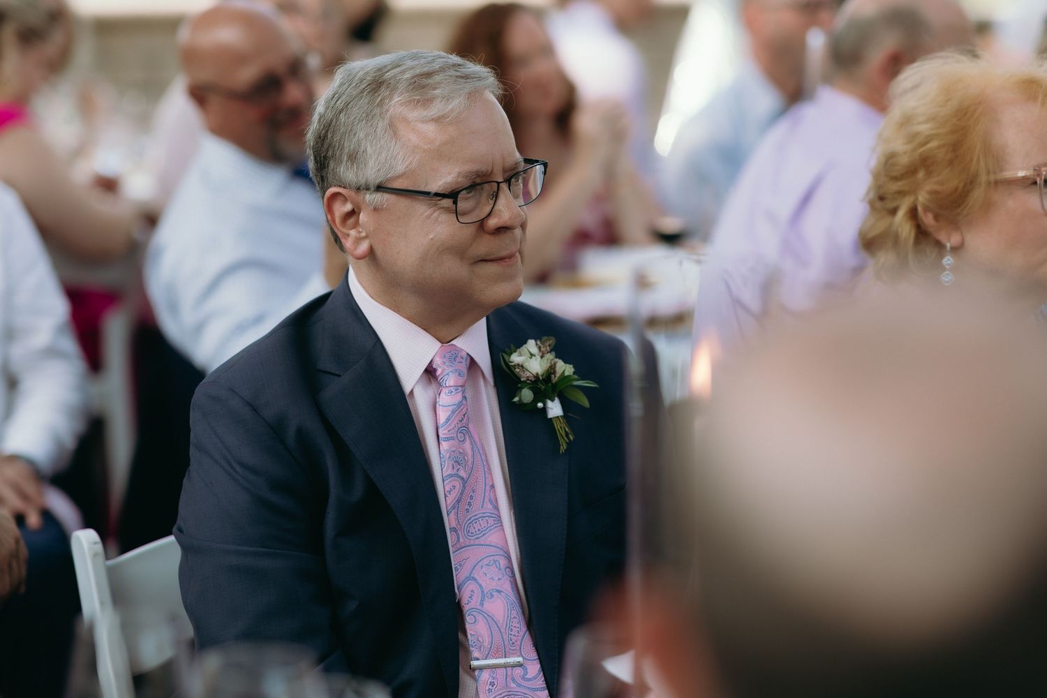 Man in navy suit and pink paisley tie wearing boutonniere seated at wedding reception.