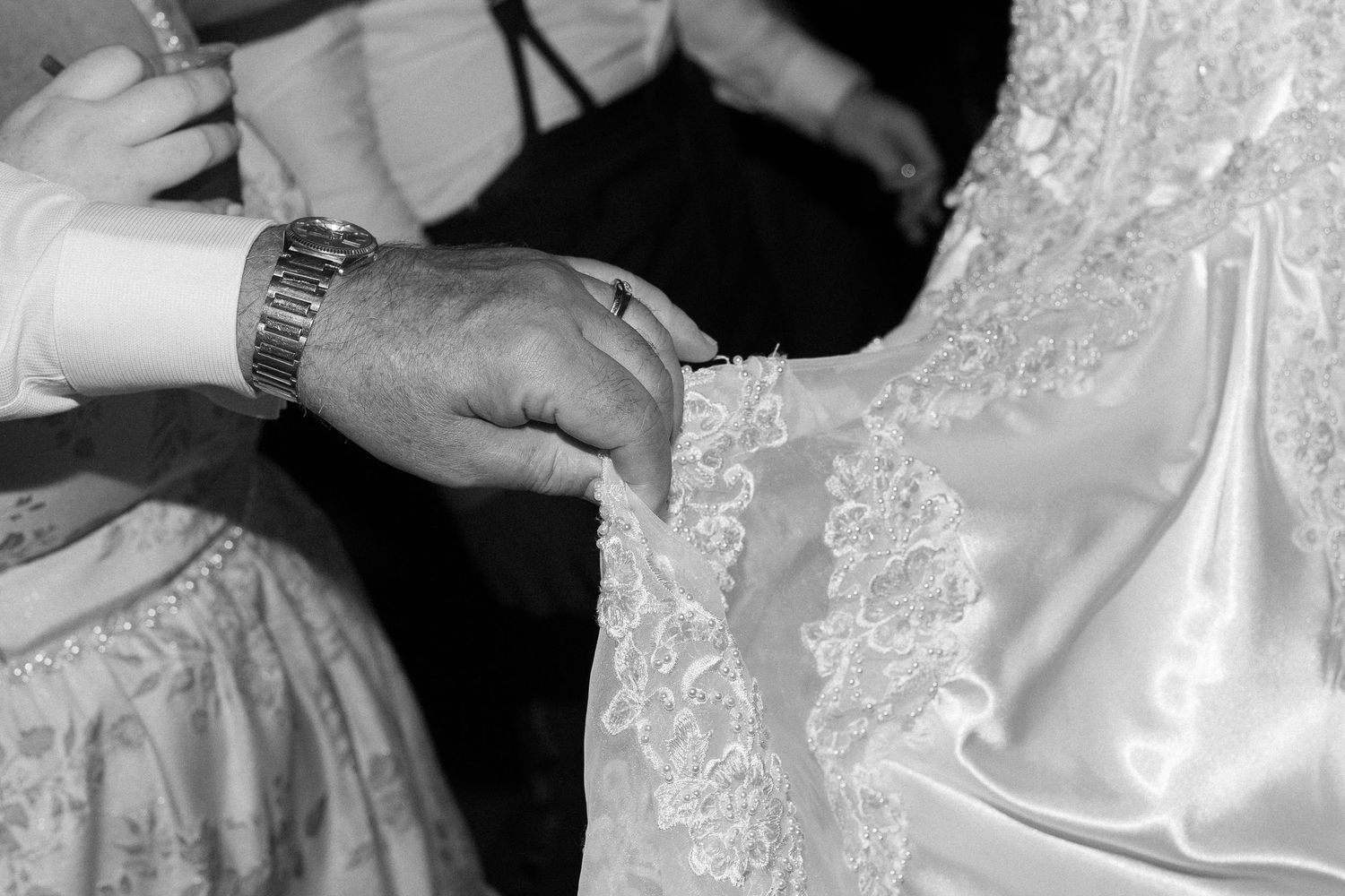 Black and white close-up of hands adjusting intricate lace wedding dress hem with watch visible.