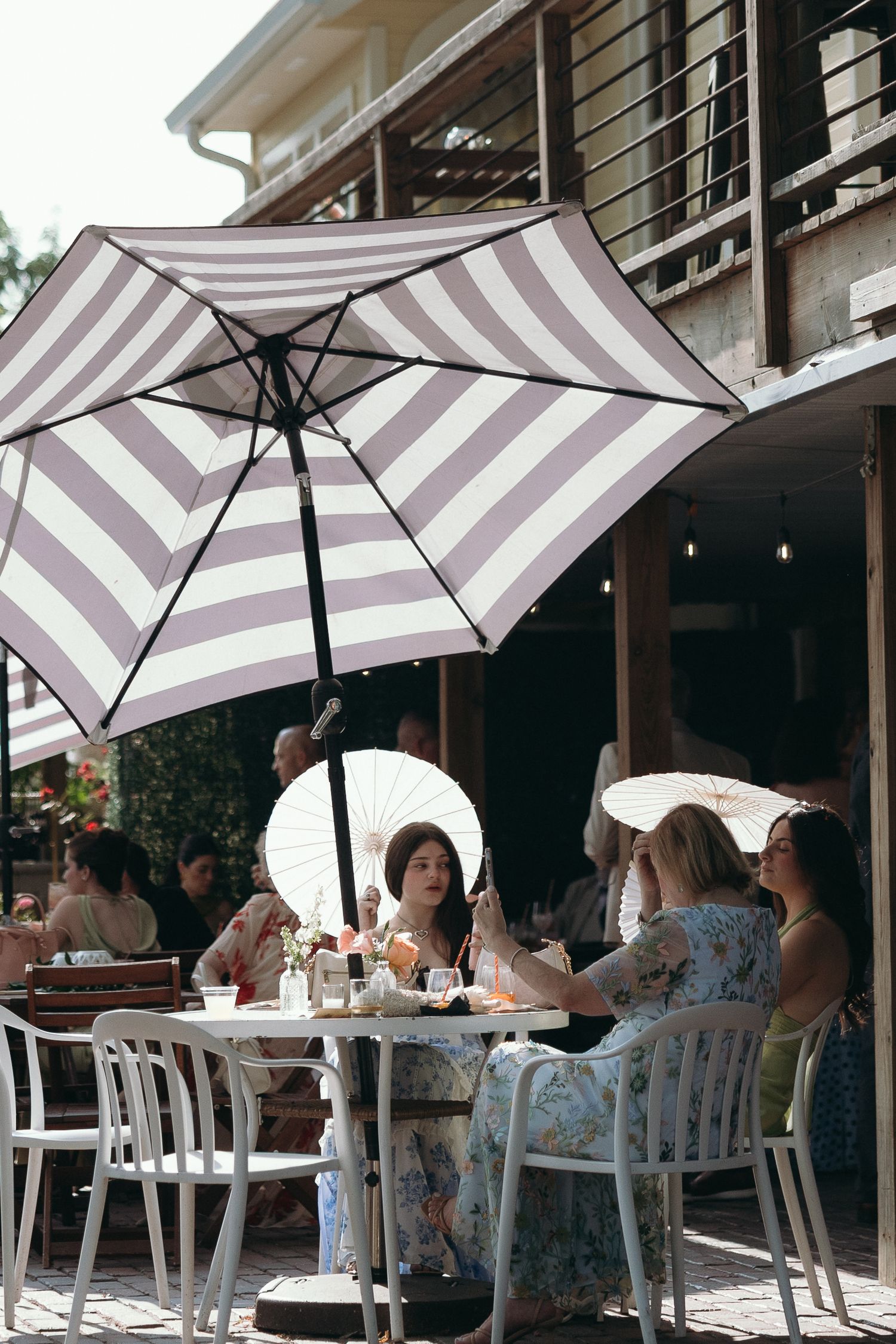 Women dining outdoors under striped purple and white patio umbrellas at a sunny restaurant terrace.