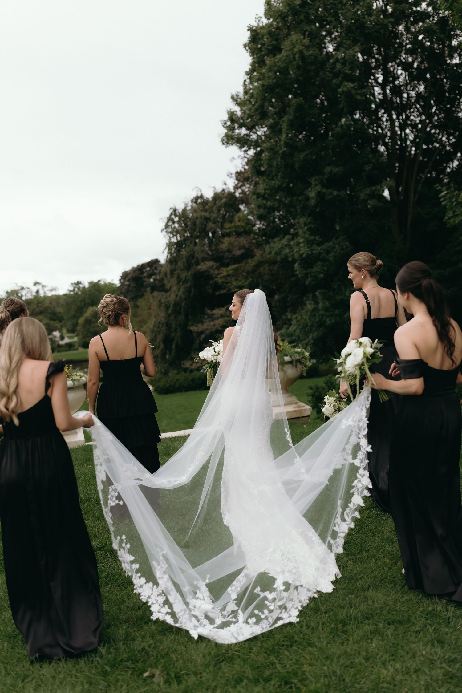 Bride in white lace veil with bridesmaids in black gowns walking outdoors on green lawn near trees.