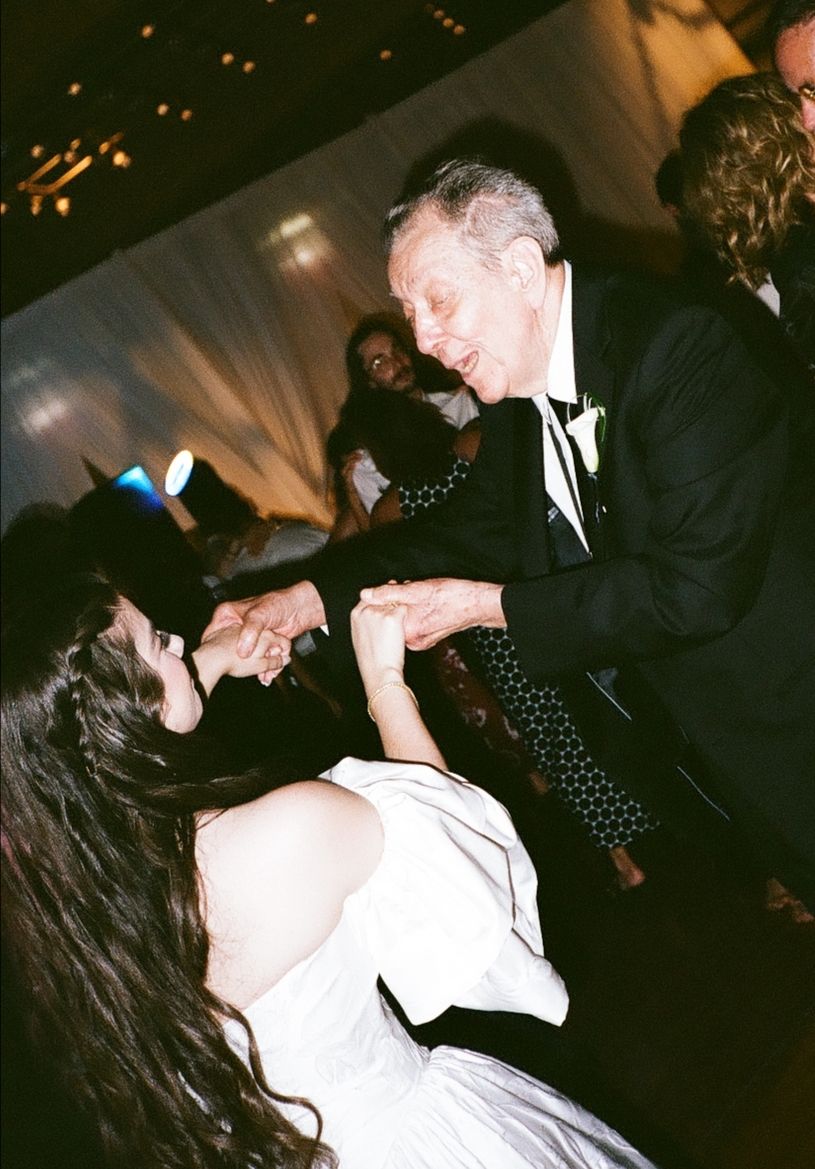 Bride and elderly man share a joyful dance at a wedding reception, holding hands on the dance floor.