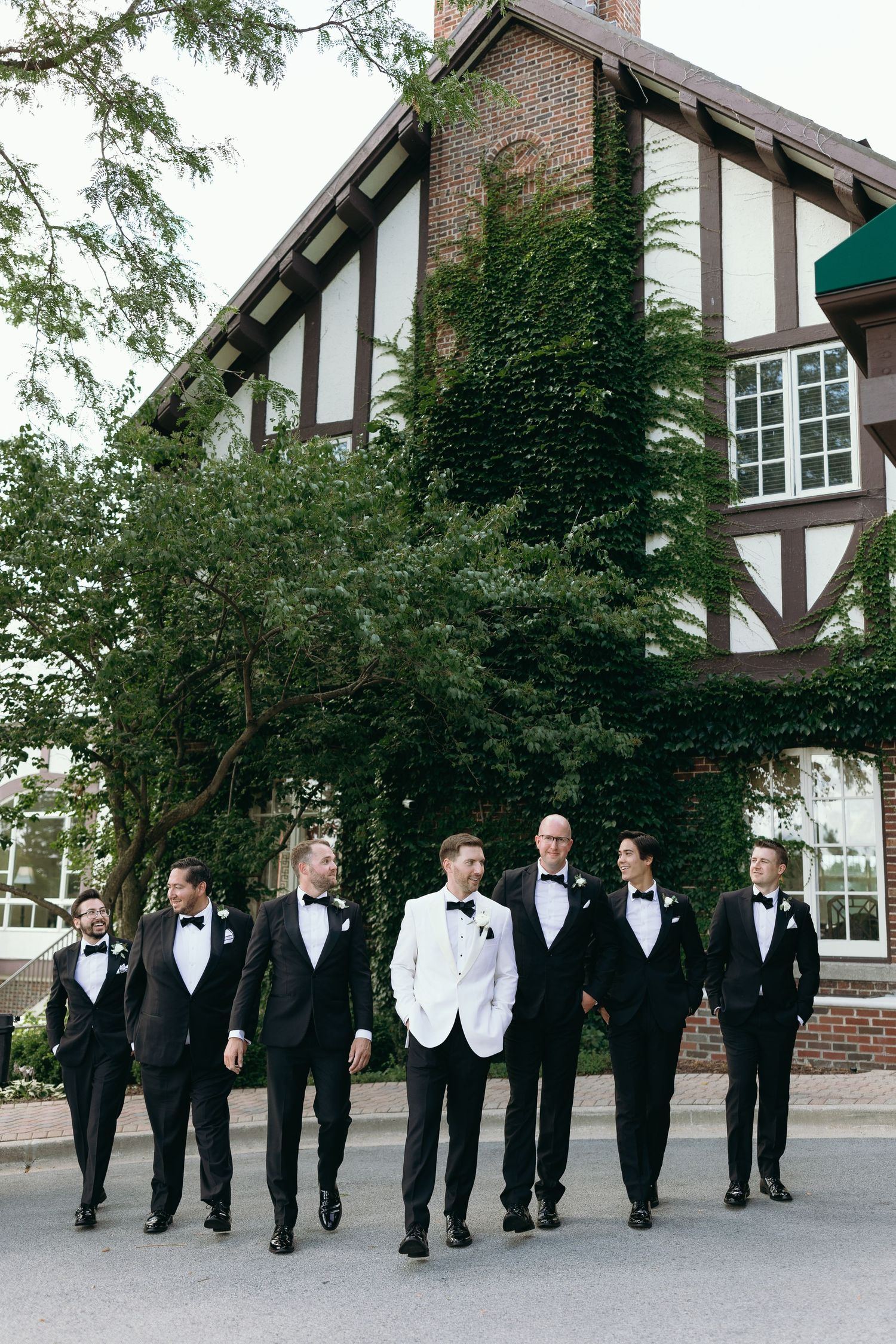 Groomsmen in tuxedos walking together outside Tudor-style building.
