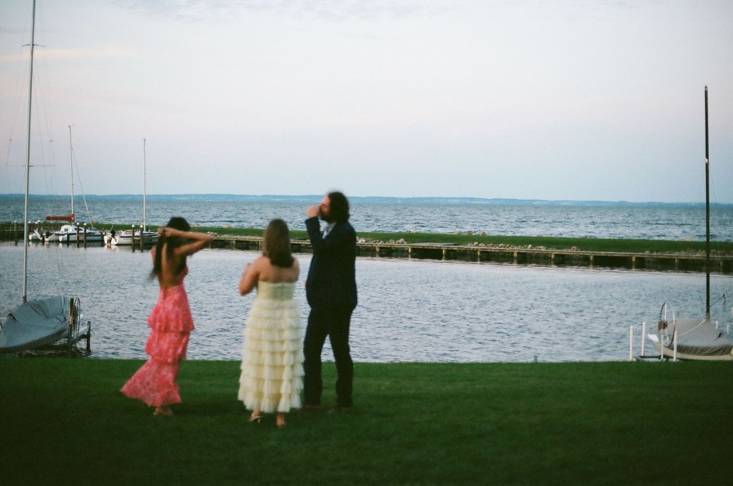 People in formal attire stand at the edge of a waterfront lawn overlooking a marina at dusk.
