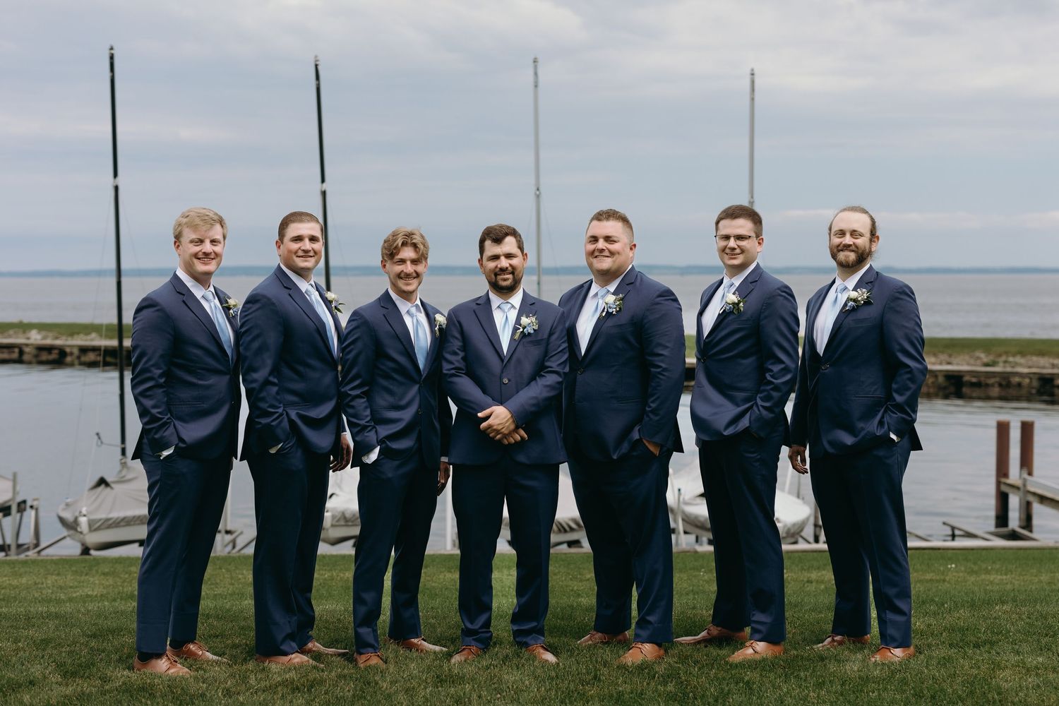 Wedding party in navy blue suits stands together on green lawn overlooking water on overcast day.
