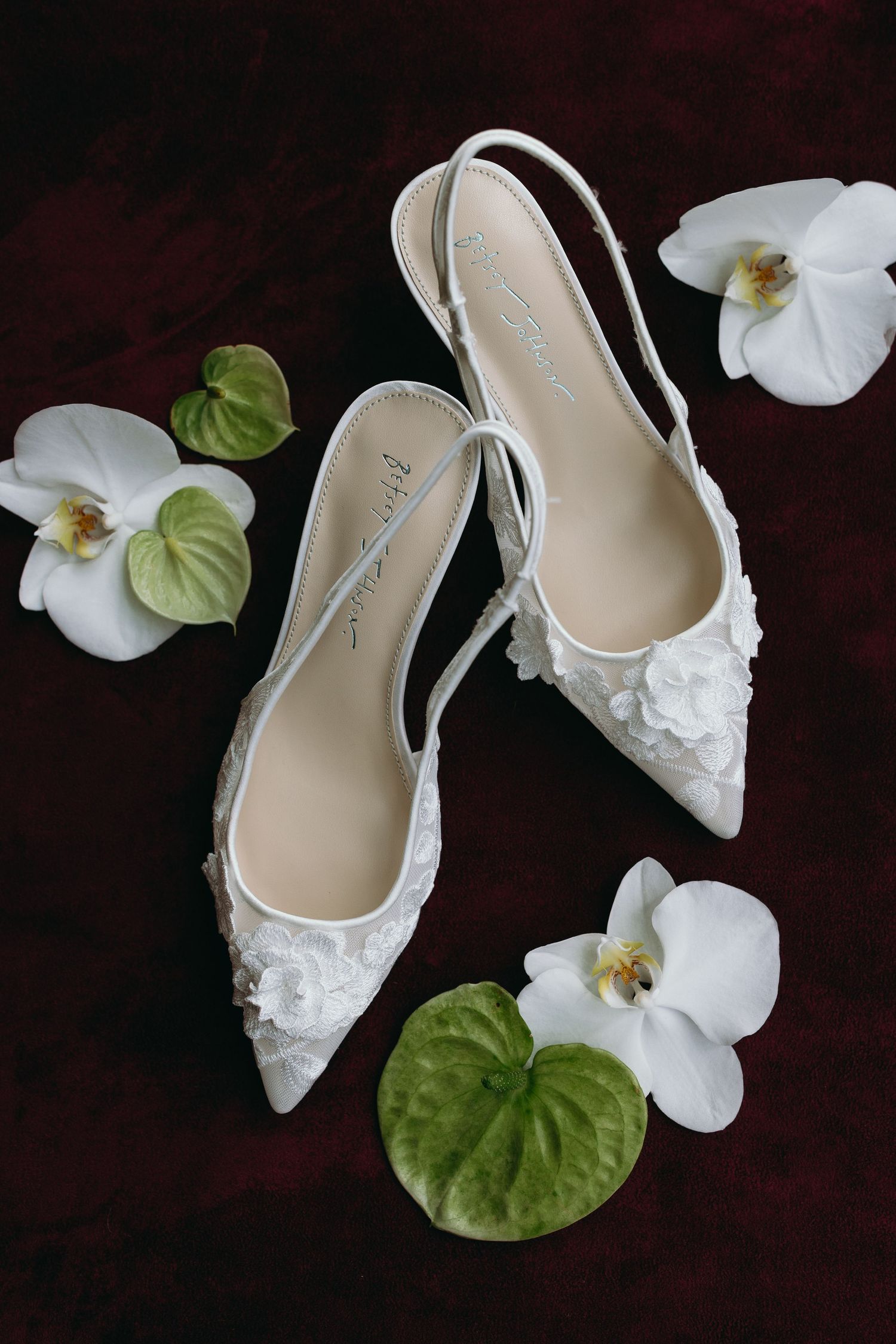 Elegant white lace bridal shoes with floral details displayed alongside delicate white orchids and green leaves.