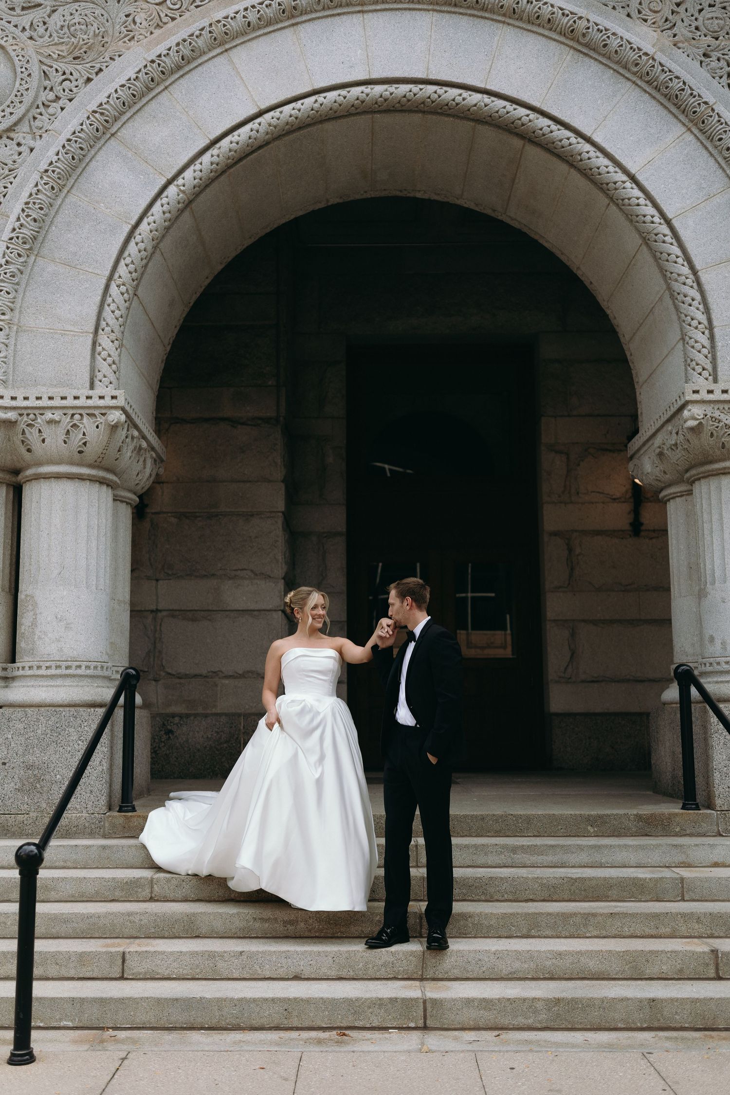A couple in wedding attire stands on stone steps beneath an ornate archway entrance of a historic building.