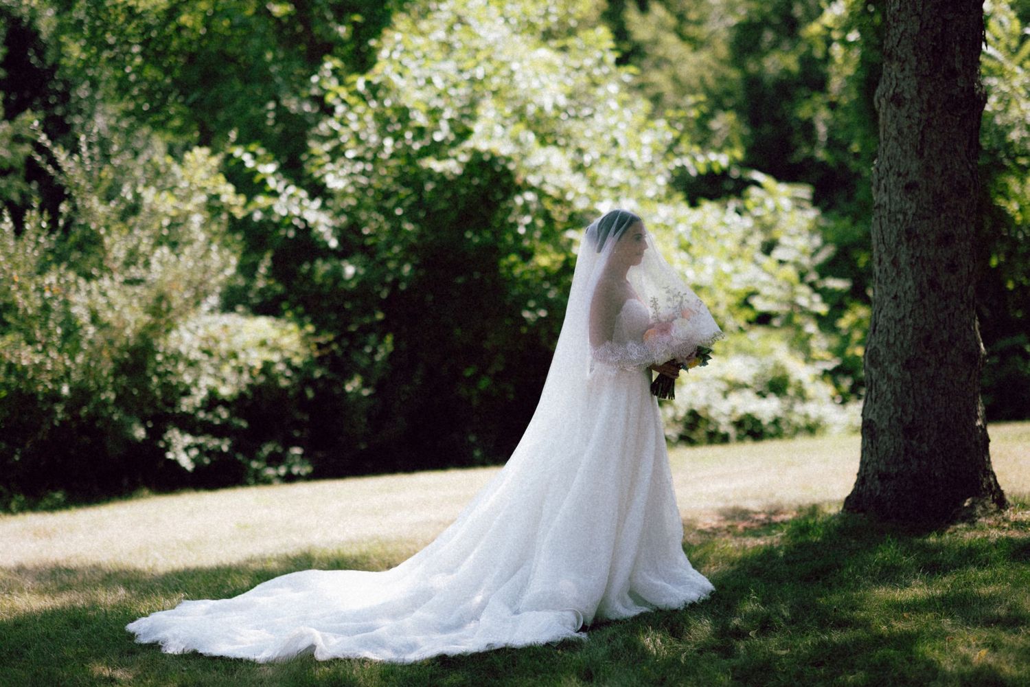 Bride in white wedding gown and veil standing near a tree in a lush green garden, holding a bouquet.