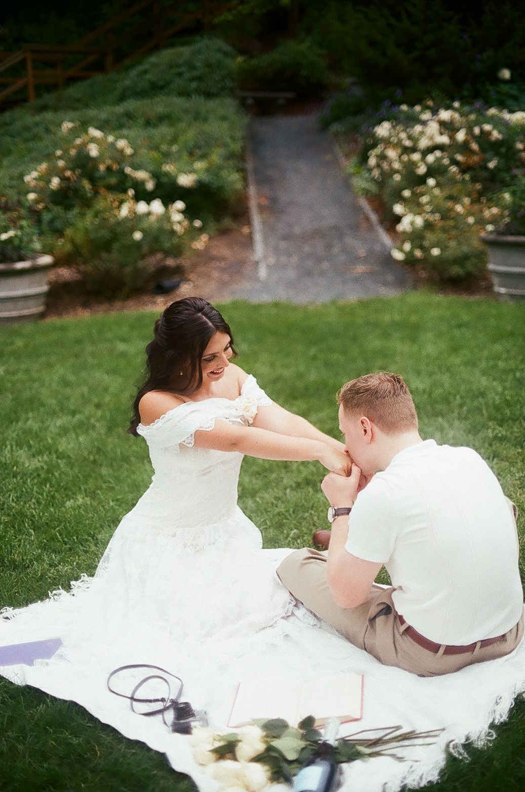 A couple sits on a white blanket in a garden with blooming flowers and a pathway in the background.