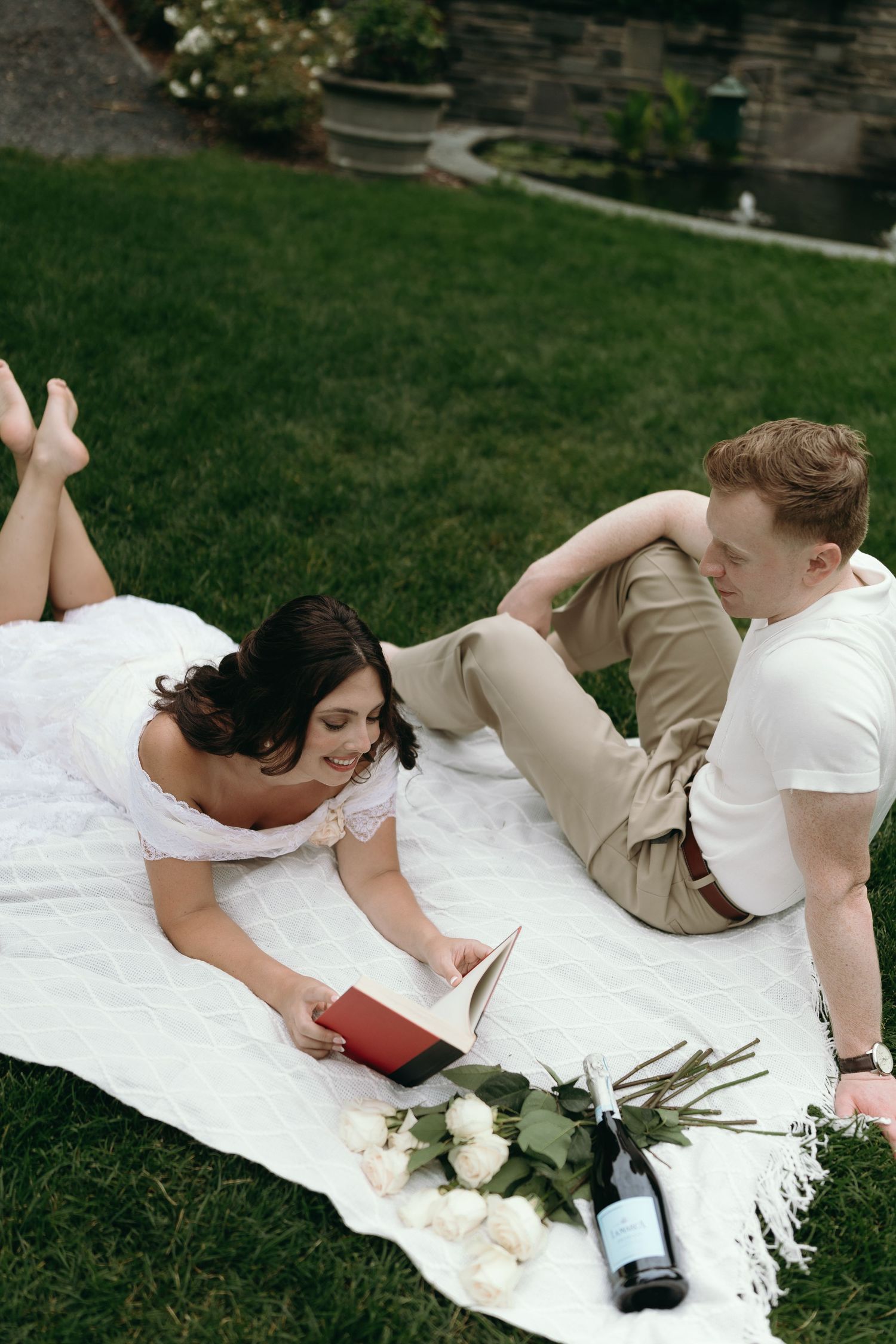 A couple lies on a white blanket on grass during an outdoor photo session with white flowers nearby.