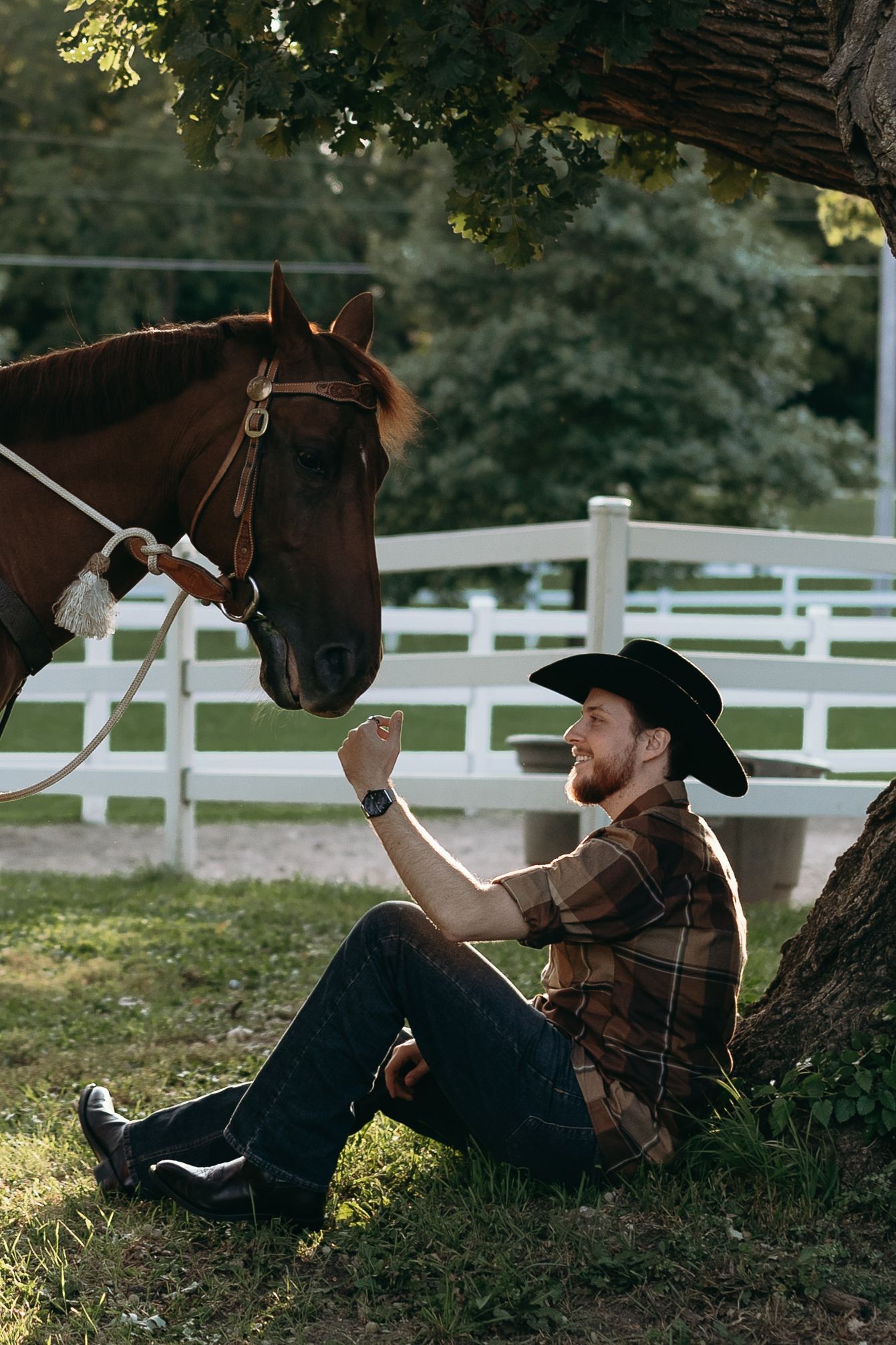 Person in western wear resting under a tree with a brown horse on a sunny day at a ranch with white fencing.